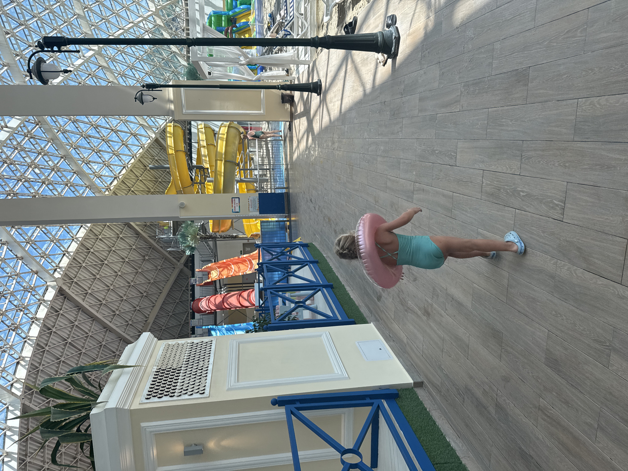 The image shows the interior of a large, indoor water park. The architecture features a high, geodesic dome roof made of glass and metal, allowing natural light to flood the space. In the foreground, a young girl with blonde hair, wearing a turquoise swimsuit and a pink inflatable ring around her waist, walks away from the camera on a wooden-tiled floor. She is also wearing blue slip-on shoes. To her left, there is a decorative wall with a plant in a planter at the top and a blue railing in front of it. In the background, several brightly colored water slides are visible, including yellow, red, and orange ones. There are also lounge chairs and cabanas in the distance, as well as other people enjoying the water park. The lighting suggests it is daytime. The location is identified as Estosadok, Russia.