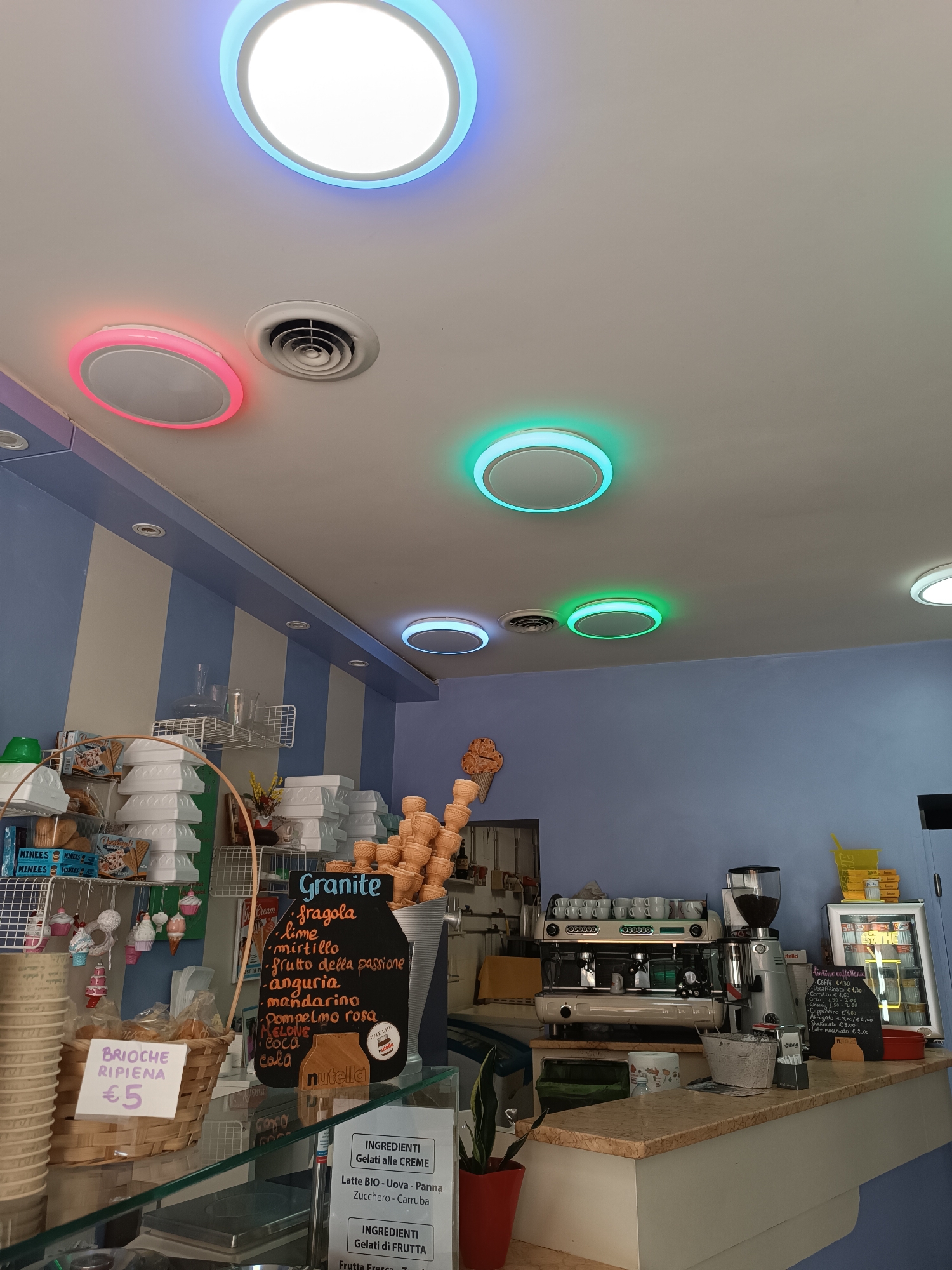 The image depicts the interior of an ice cream shop. In the foreground, a glass counter displays various items, including stacks of paper cups, a basket with a sign reading "BRIOCHE RIPIENA €5", and a menu board listing ice cream flavors and prices. Behind the counter, an espresso machine and coffee grinder are visible, along with stacks of ice cream cones. The walls are painted a light blue and cream color, and the ceiling features circular lights with colored rims in blue, pink, and green. The overall impression is a well-lit and organized retail space. The location is identified as Casalmaggiore, Italy. There are no people visible in the image.