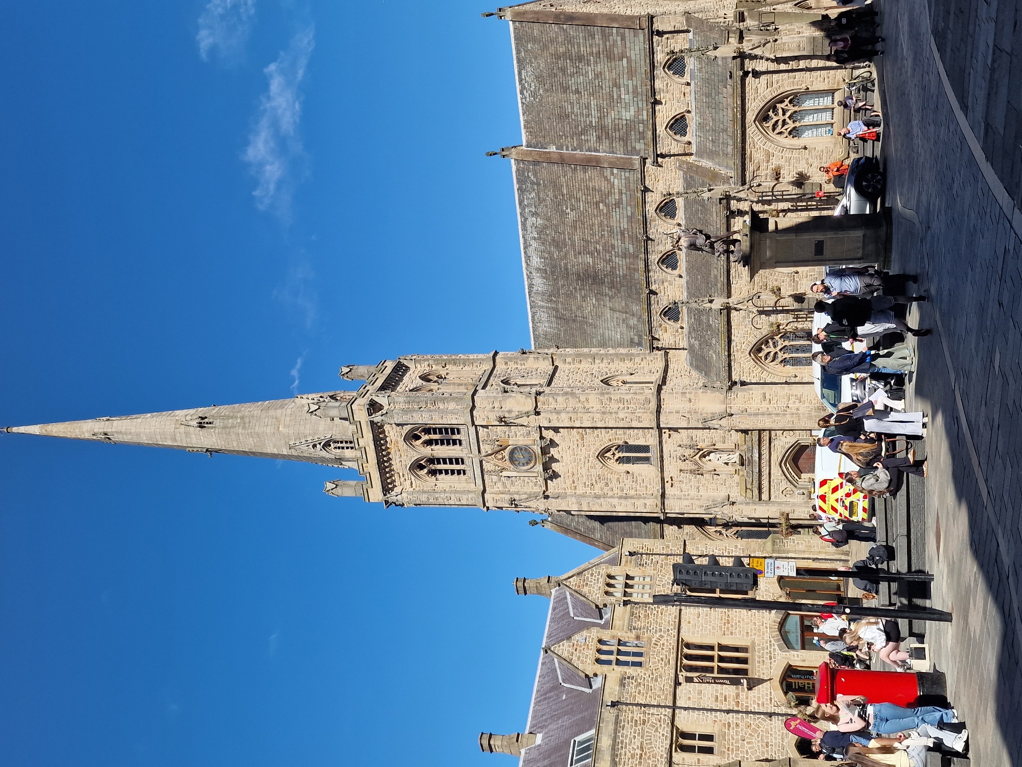 The image captures a scene in Durham, United Kingdom, featuring the Durham Cathedral spire against a bright blue sky with some wispy clouds. The architecture of the cathedral is visible in detail, including the stonework, windows, and various decorative elements. The image shows the cathedral facade and nearby Town Hall.
The street in front of the building is populated with pedestrians. A red postbox is near people sitting and lying on the steps in front of the Town Hall. A white van with reflective markings is parked on the road near a traffic signal. Groups of people are gathered in various spots, some standing, others sitting on steps or benches.