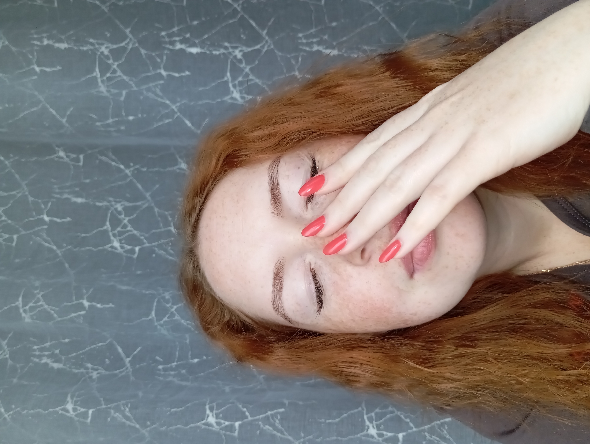 A person with red hair and freckles is lying down on a grey surface with a marble-like pattern. Their eyes are closed, and their hand is covering their face. The person is wearing red nail polish. The setting appears to be indoors. The city of Yurgamysh, Russia, is not discernable from the image.