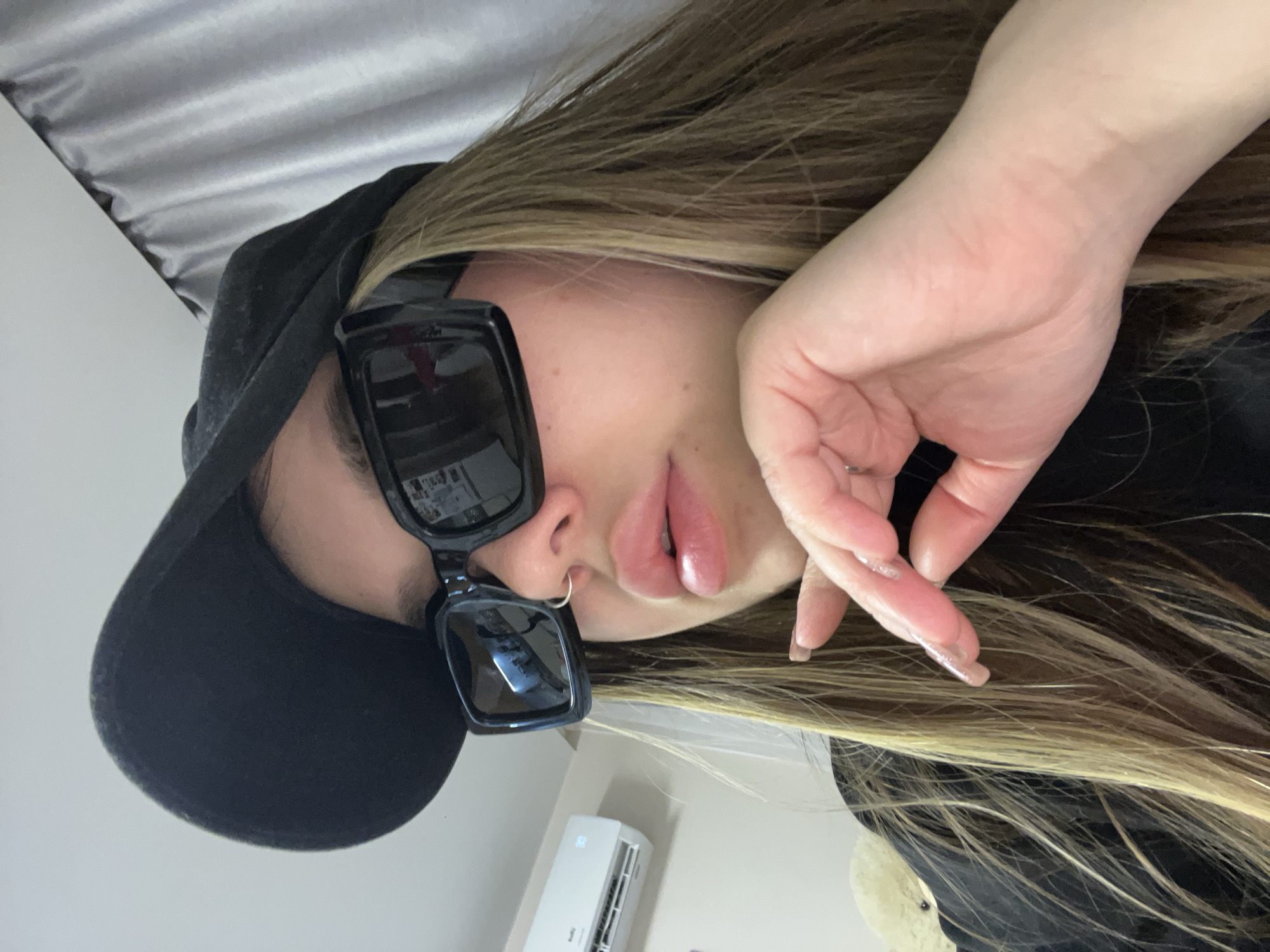 A close-up, portrait-style photograph, taken with the subject lying on their side, captures a person, likely female, indoors. The individual wears large black rectangular sunglasses and a black baseball cap. Long, straight hair, appearing to be a blend of blonde and light brown, frames her face. Her full lips are visible, and she has a small silver hoop nose ring. One hand, featuring long, manicured nails with a clear or light polish, is raised towards her face in a relaxed pose.
In the background, a light-colored wall is visible, with a white air conditioner unit mounted on it, displaying the number "26" on its digital screen. Above the subject's head, a textured grey and white surface, possibly a curtain or a part of the ceiling, adds to the indoor setting. The image is taken in an indoor environment, with no discernible outdoor elements or time of day. The context for the image is Krasnodar, Russia.