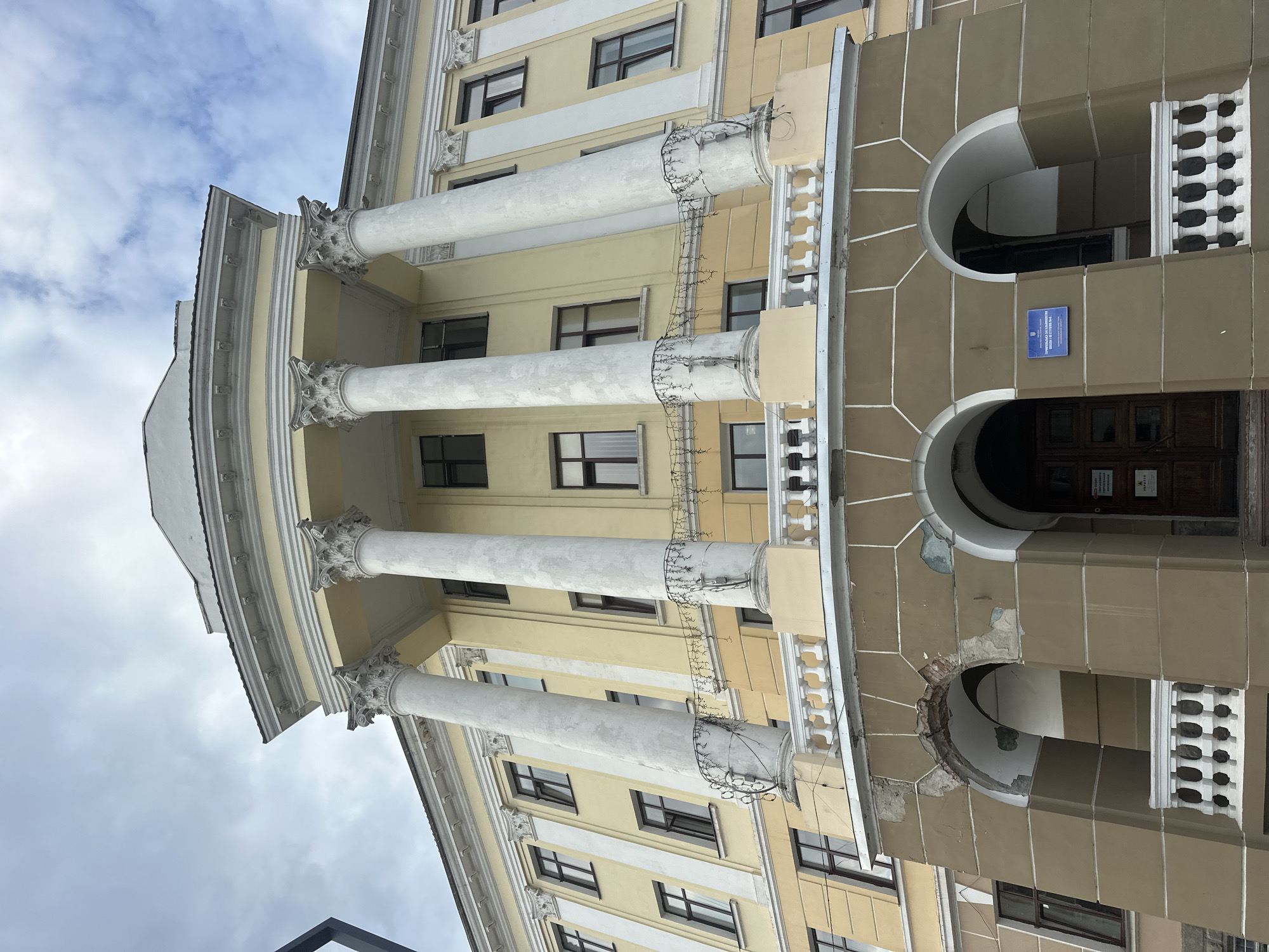 The image shows the facade of a light yellow building with white columns and architectural details. The building has a semicircular portico supported by large, fluted white columns. Above the portico, there is a balcony with a white balustrade. The lower part of the building features arched entrances. One of the arches shows damage to the plaster. A sign with blue and white text is visible on the facade above one of the entrances. The sky is cloudy. This building is located at 2 Ternopilsky National Economic University Street in Ternopil, Ukraine, and it is the main building of the Ternopil National Economic University.