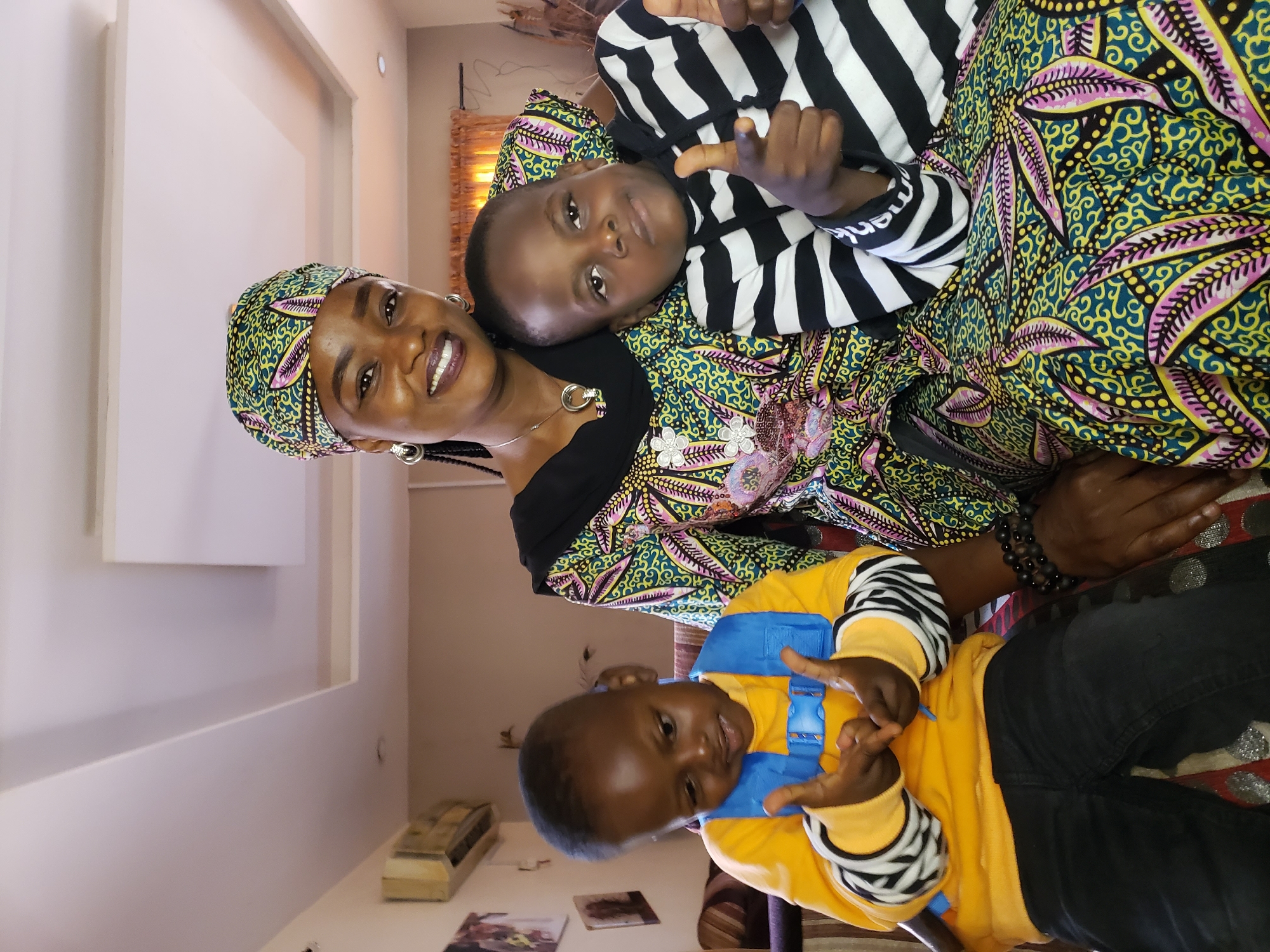 In Sabon Tasha, Nigeria, a woman with dark skin, wearing a vibrantly patterned headscarf and dress, smiles at the camera. She is seated, with a younger child on her left and an older child on her right. The younger child, wearing a yellow long-sleeved shirt and black pants with a blue harness, looks down with hands raised. The older child, in a black and white striped long-sleeved shirt, gives a thumbs-up gesture towards the camera. The environment appears to be an indoor setting, possibly a living room, with a patterned sofa and ceiling lights visible.