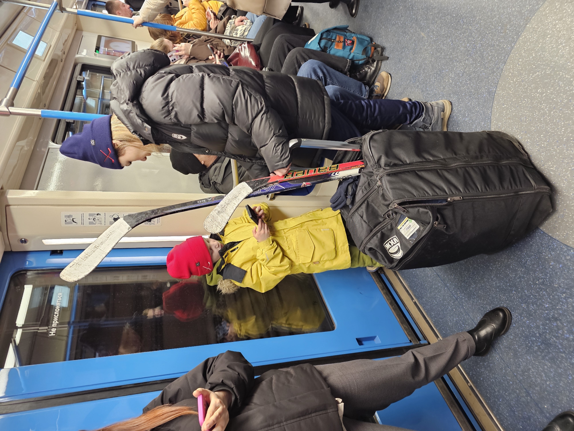 The image captures a scene inside a subway train car, oriented vertically. Several passengers are visible, mostly seated or standing.
The main subjects are a young person with blonde hair wearing a dark blue beanie with a red "X" design and a black puffer jacket, and a child in a red beanie and a bright yellow puffer jacket. The blonde-haired individual is standing, leaning against a grab bar, and holding two hockey sticks with one hand. One stick has "Bauer" visible on it. The child is seated on what appears to be a bench next to a large black hockey equipment bag, looking down at a mobile phone. The hockey sticks are partially inside the equipment bag.
Other people in the train include individuals seated in the background, one person holding a pink phone with red hair partially visible in the foreground, and various legs and feet throughout the car. One person's legs and feet are visible in the lower right, wearing grey trousers and black dress shoes. Another's legs wear jeans and brown/grey shoes, next to a blue and green backpack.
The setting is clearly a subway train, indicated by the blue grab bars running along the ceiling, the typical seating arrangement, and the sliding doors. The floor has a speckled blue pattern. The attire of the individuals (puffer jackets, beanies) suggests cold weather outside.
Visible text includes "Не прислоняться" (Do not lean / Do not lean against) on the blue train door, indicating a Russian-speaking environment. On the black hockey bag, some text appears, possibly including "JXXA" and "Охрана" (Security), though it is partially obscured and not fully legible. The overall activity is people commuting by subway, with the two central figures traveling with sports equipment.