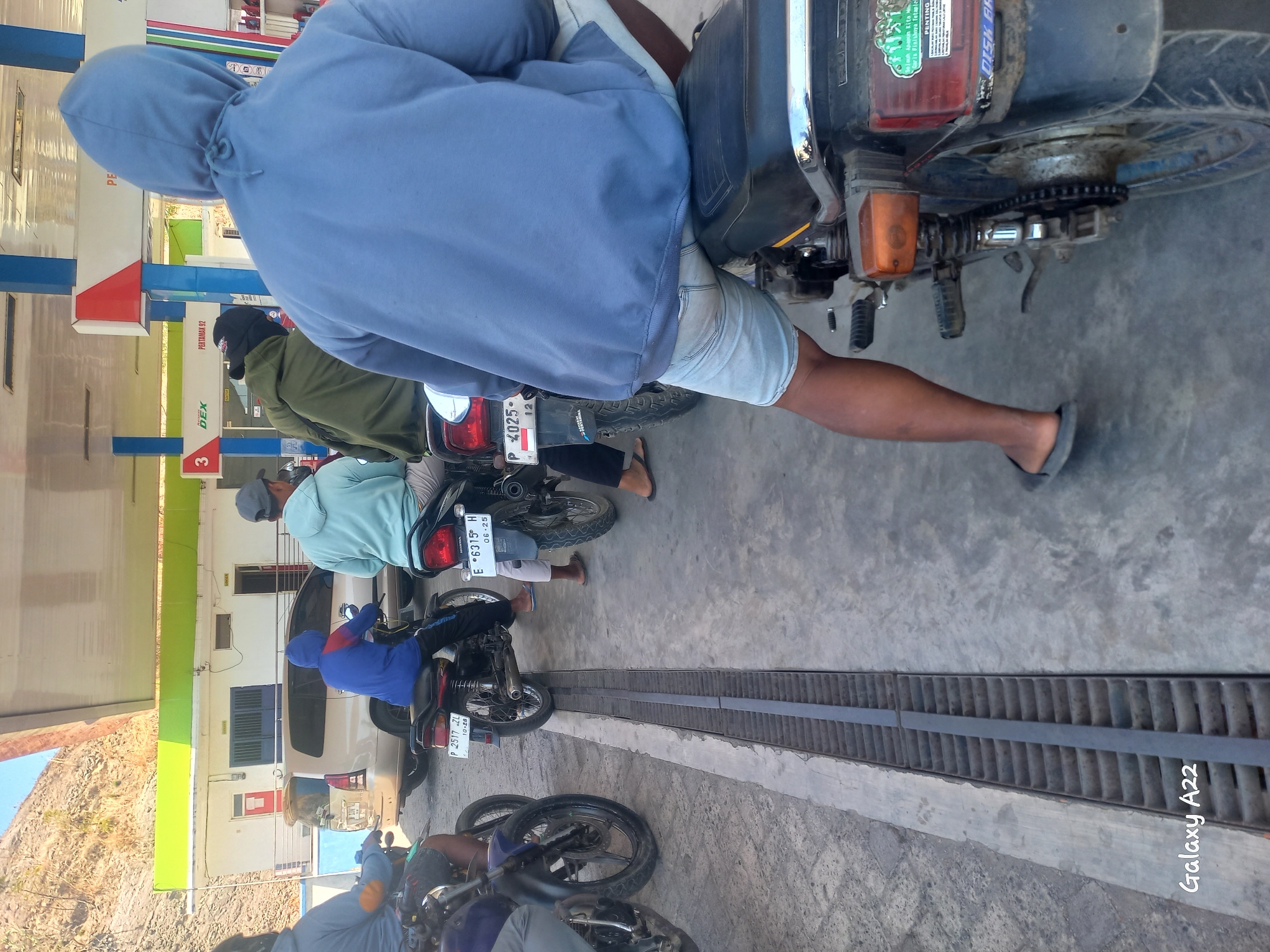 At a Pertamina gas station in Kotakan, Indonesia, a line of motorcyclists waits for fuel. The scene is captured on a sunny day, with a mountainous backdrop visible in the distance. A white building with green trim and blue pillars stands behind the gas pumps, which are partially obscured.
Several individuals are seated on their motorcycles, facing away from the camera. One person in the foreground wears a blue hoodie and light blue shorts. To their left, a motorcyclist in a blue jacket and black pants is also visible. Further back, another person in a light green hoodie and a person in a dark green jacket are waiting in line. A light-colored van is parked behind the first few motorcycles.
The ground is paved with concrete and features a metal grate drain running parallel to the motorcycles. The motorcycles themselves appear to be older models, with visible license plates and exhaust pipes. The overall impression is one of a typical, bustling gas station scene in a moderately populated area.