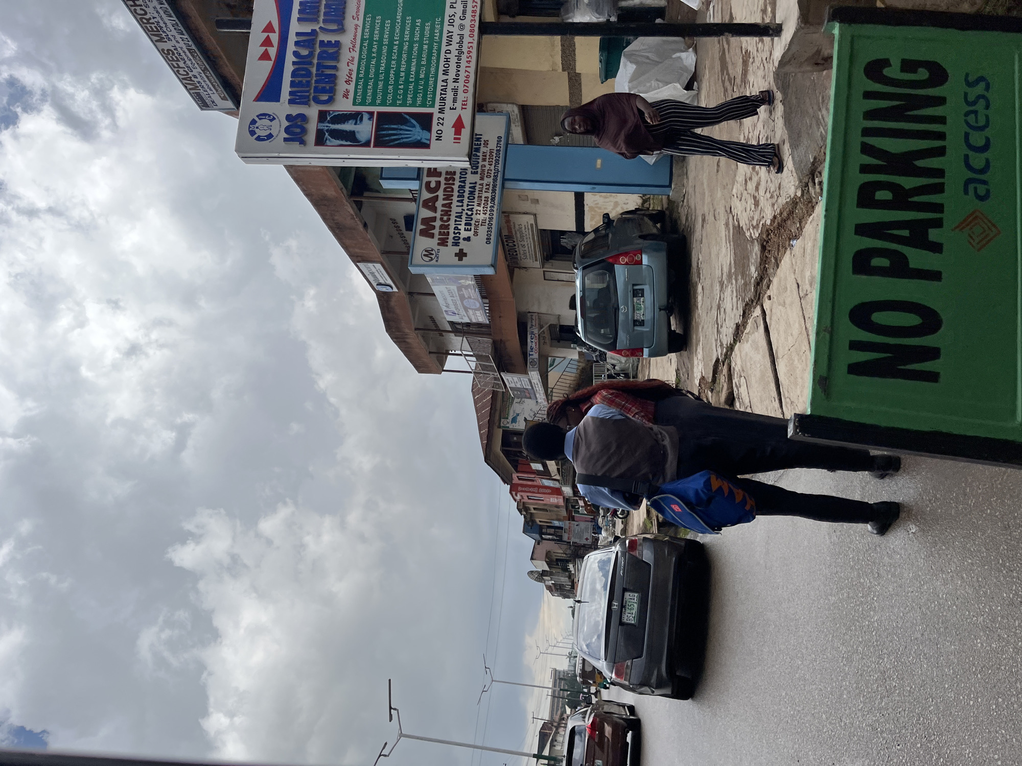 This is a medium-angle, eye-level shot of a street scene in Jos, Nigeria. The perspective is rotated, presenting the image sideways. The sky is overcast with thick clouds, suggesting a dull or possibly rainy day.
The foreground features a street with several cars, including a grey Honda sedan with the license plate "BSA-651AG." A prominent green sign with "NO PARKING" and the "access" logo is on the right side of the frame.
In the middle ground, a group of people are walking. Two individuals appear to be carrying a blue bag, suggesting they are either traveling or moving goods. One person is wearing a dark headscarf and striped pants. Another person is wearing a grey vest over a blue shirt.
The background reveals a row of buildings lining the street, characteristic of a commercial area. Several signs are visible on these buildings, including one for "JOS MEDICAL IMPERIAL CENTRE" which lists various medical services like "GENERAL RADIOLOGICAL SERVICES" and "ROUTINE ULTRASOUND SERVICES." Another sign indicates "MACFES MERCHANDISE HOSPITAL, LABORATORY & EDUCATIONAL EQUIPMENT." The presence of these businesses suggests a busy urban environment. The overall scene depicts a moment of daily life on a Nigerian street.