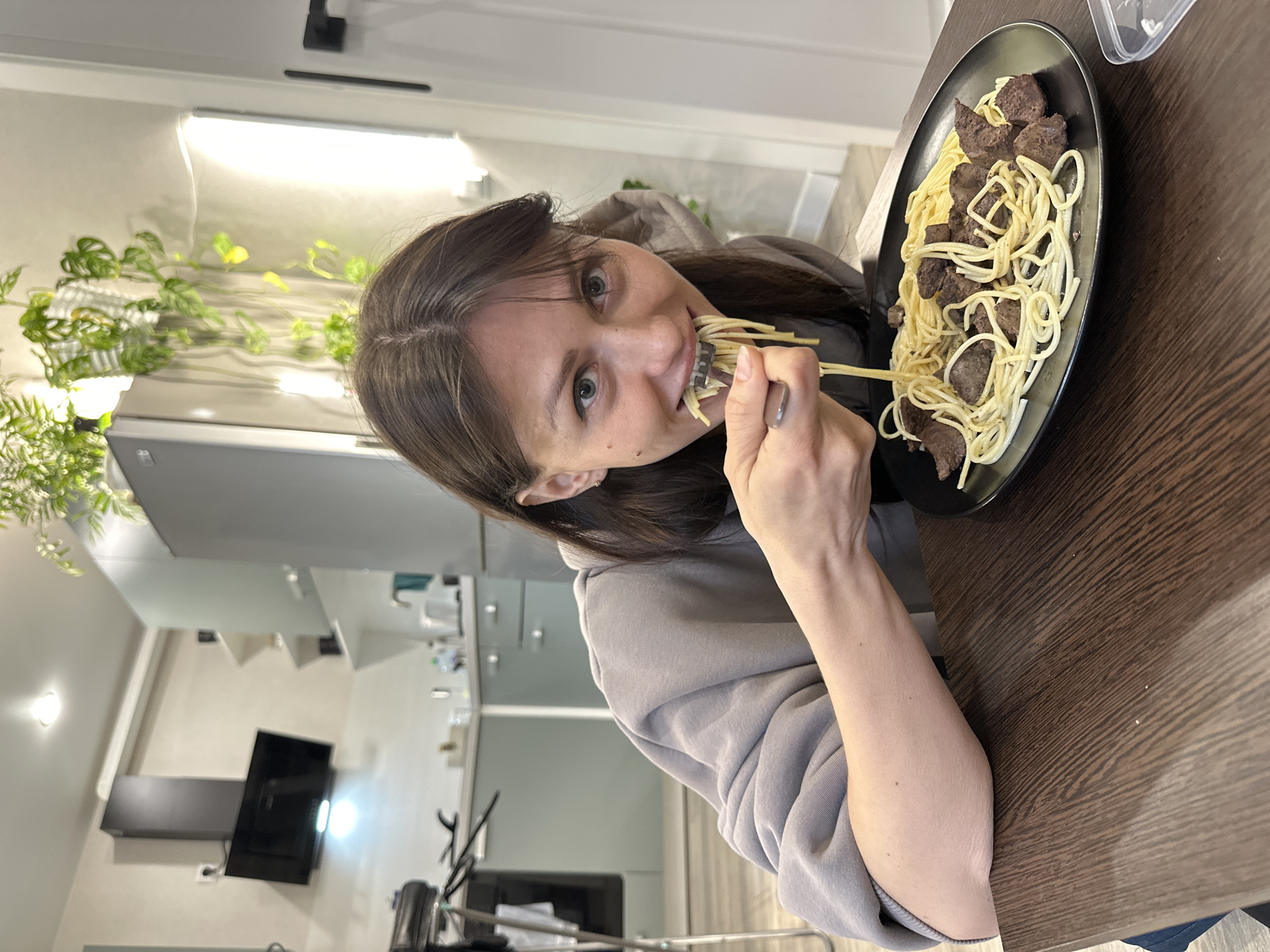 A young woman is captured mid-meal in an indoor setting, likely a home kitchen or dining area in Murino, Russia. She has dark brown hair parted in the middle and light-colored eyes, looking directly at the camera with a playful or engaged expression as she takes a bite of spaghetti from a fork. Her mouth is partially open, containing strands of pasta, and her right hand, holding the fork, is prominent. She is wearing a light grey hooded sweatshirt.
On a dark brown wooden table in front of her, a black oval plate holds a serving of spaghetti mixed with several dark brown, cubed pieces of meat, possibly liver or stew. A clear plastic container is visible on the table to the right of the plate.
The background reveals a modern interior. To her left, several green houseplants are arranged, illuminated by horizontal white LED strip lights, possibly serving as grow lights. Further back, blurred kitchen appliances such as a refrigerator, a black range hood, and a cooktop are visible. A black door handle can be seen in the upper left corner. The scene conveys a casual, everyday moment, with the interior lighting suggesting an indoor shot where artificial light supplements natural light for the plants.