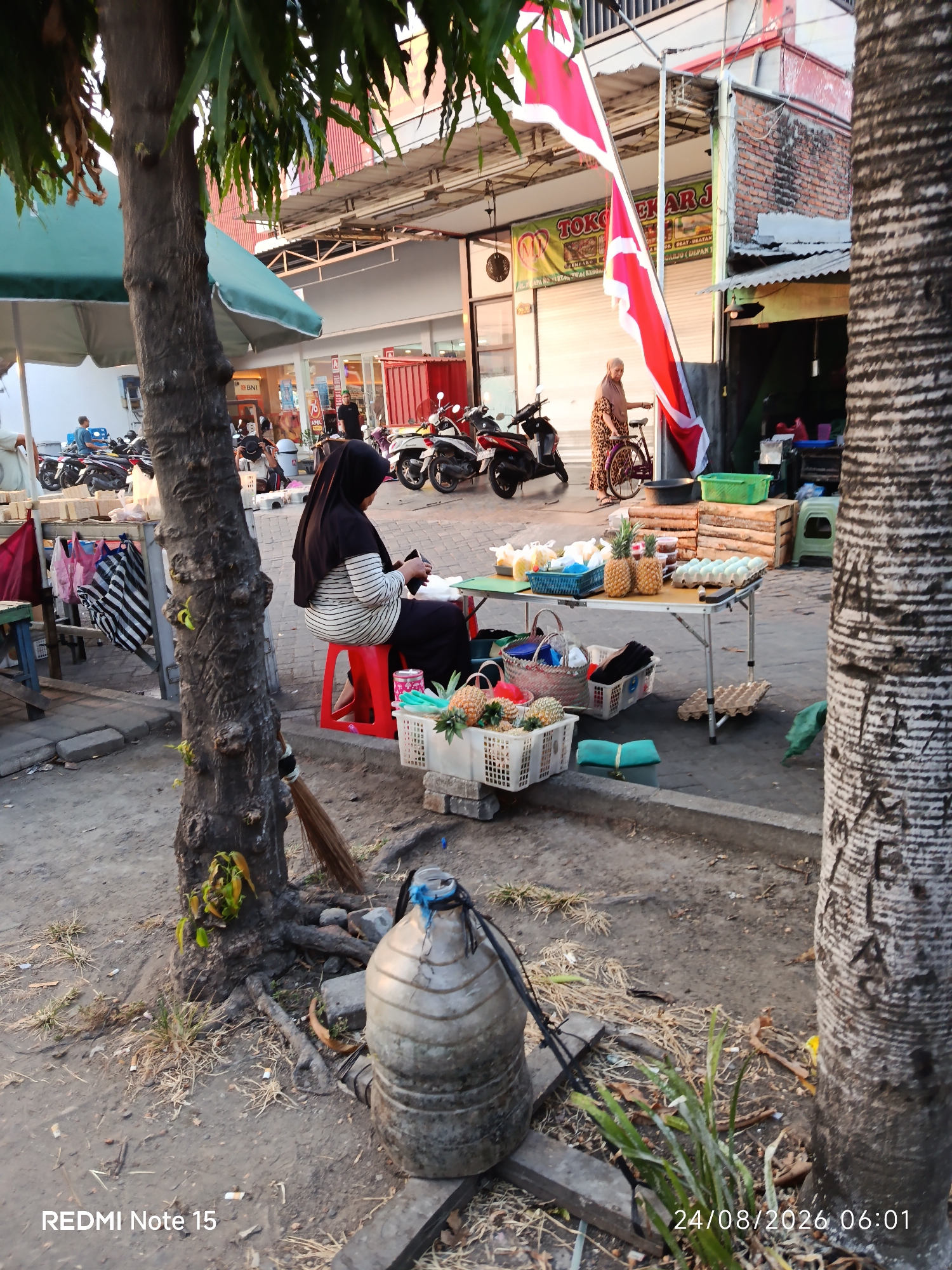 The image depicts a street scene in Keboansikep, Indonesia, likely a marketplace or roadside stall area. A woman wearing a black hijab and a striped shirt is seated on a red stool, looking at her phone. In front of her, a table displays various items including pineapples, baskets of produce, and a carton of eggs. To the left, a tree trunk is prominent, with foliage overhead and a green umbrella partially visible. In the background, several motorcycles are parked, and a building with a red and brick facade suggests a commercial area. A red and white flag is also visible. The ground is dirt and pavement, with a large plastic water jug in the foreground. The timestamp "24/08/2026 06:01" and "REDMI Note 15" are superimposed on the image.