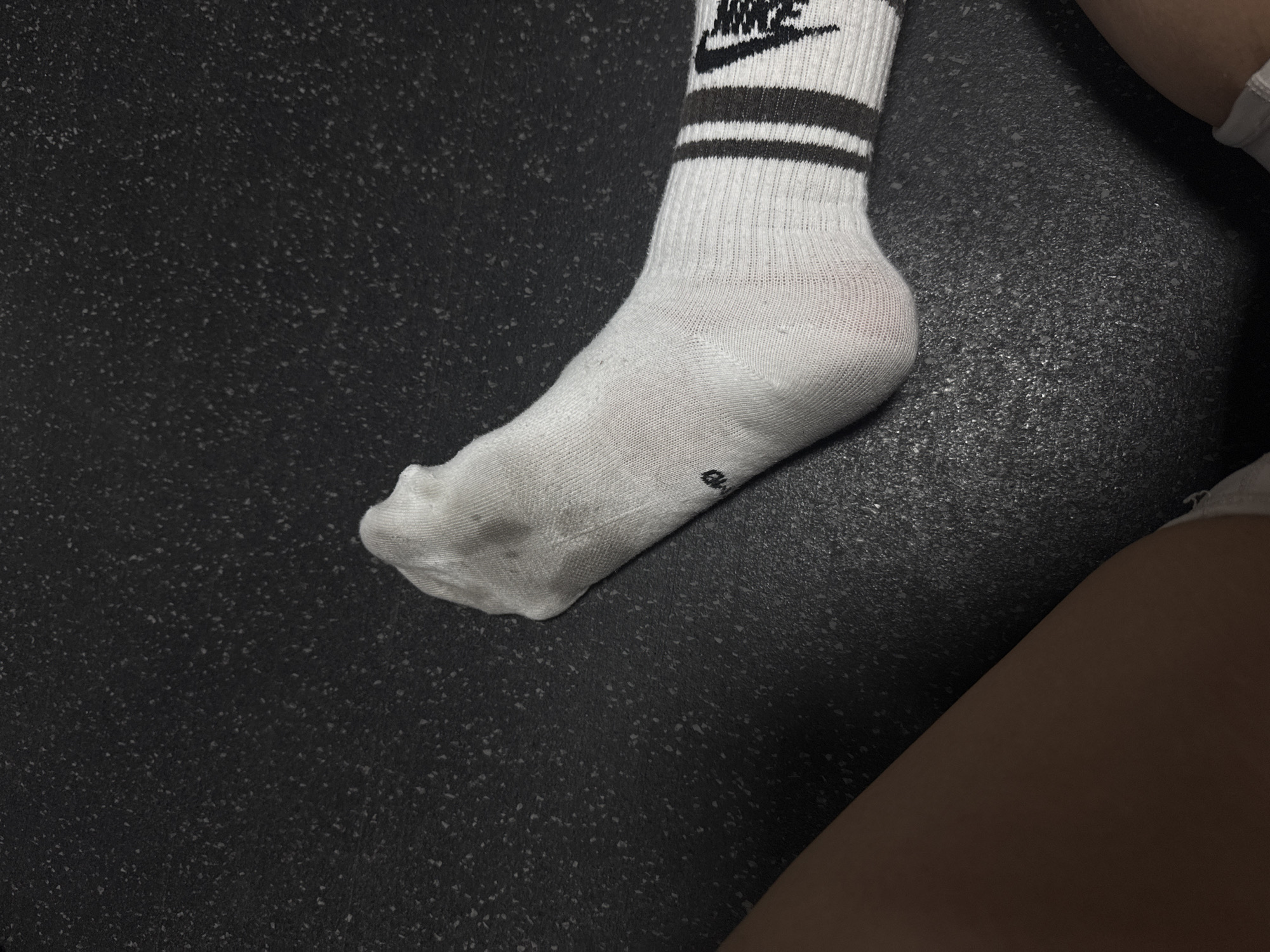 The image shows a person's foot wearing a white Nike sock with black stripes and a hole in the toe. The foot is on a dark, textured surface. Part of a leg is visible in the lower right corner. The setting is not identifiable as Dubai, United Arab Emirates, as there are no contextual cues to determine the location.