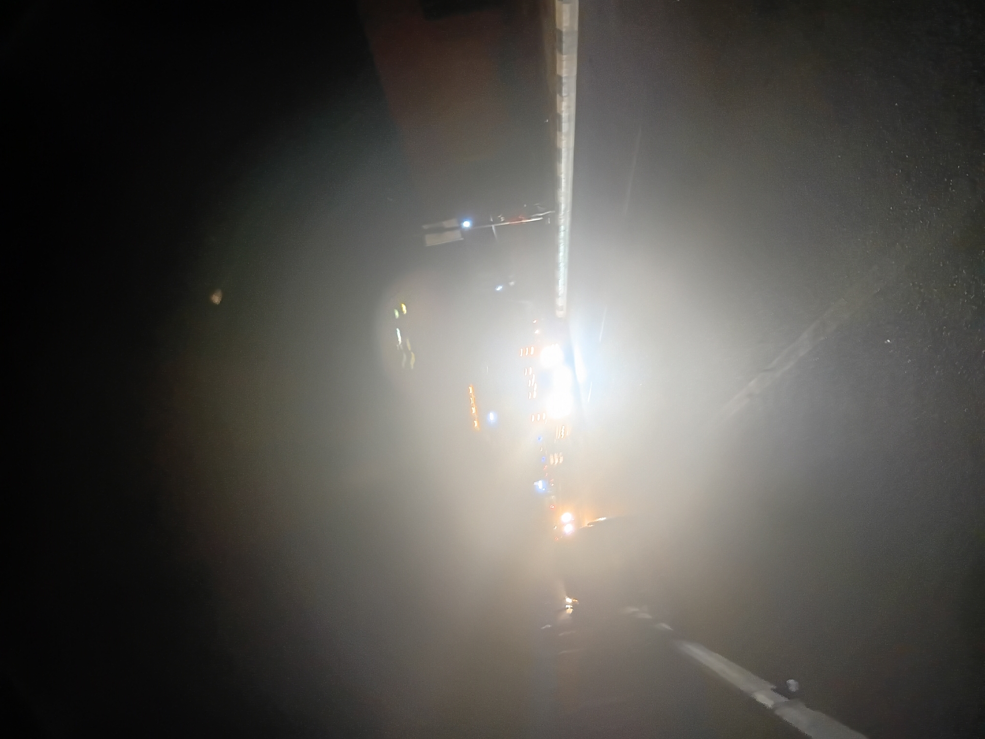 The image captures a road scene at night, likely in Jalingo, Nigeria, characterized by heavy fog or mist that significantly reduces visibility. A vehicle with bright headlights, possibly a truck or bus due to its size and the row of orange lights visible on its top, is the most prominent subject, approaching from the distance. Its headlights create a strong glare that diffuses through the fog. Other vehicle lights, appearing as blue and orange blurs, are also visible, suggesting other traffic. The road surface is dark and wet, with white lines marking the lanes. To the right, a concrete barrier with reflective strips runs along the side of the road, and a lamppost or traffic signal is dimly visible. The overall atmosphere is obscured and somewhat eerie due to the atmospheric conditions.