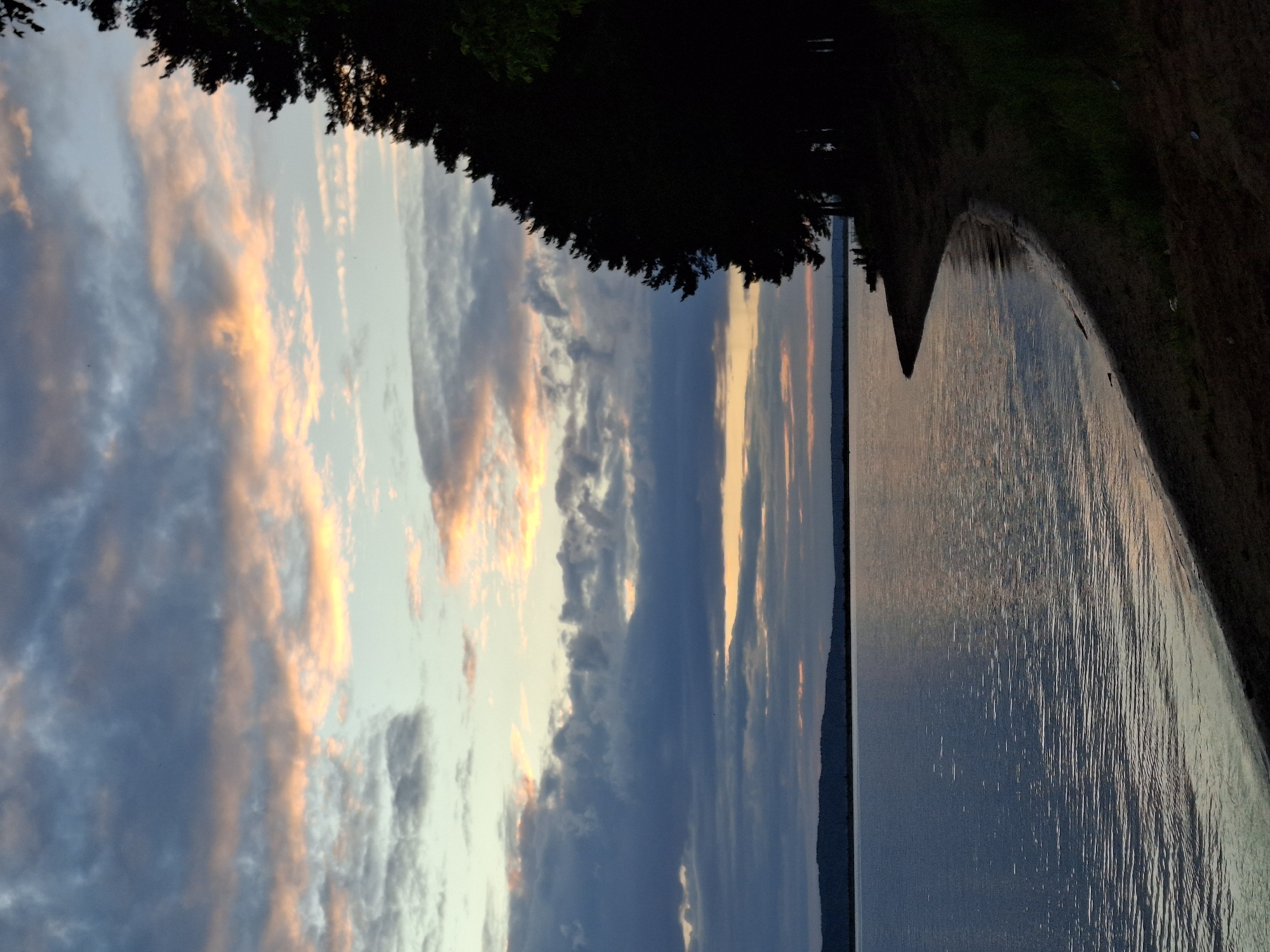 The image depicts a twilight scene by a large body of water, likely the Kama River, near the city of Chaikovsky, Russia. A dark, silhouetted line of trees runs along the right side of the frame, meeting a sandy shore that curves into the water. The water's surface is calm, reflecting the muted colors of the sky. The sky itself is filled with dramatic clouds, tinged with shades of gray, blue, and soft orange and pink hues, suggesting the last moments of daylight. In the distance, a low, dark horizon line indicates a further shore or landmass. A few small figures are visible on the distant shore, silhouetted against the water, suggesting human presence but offering no details about their actions. The overall atmosphere is serene and tranquil, characteristic of a lakeside sunset or sunrise.