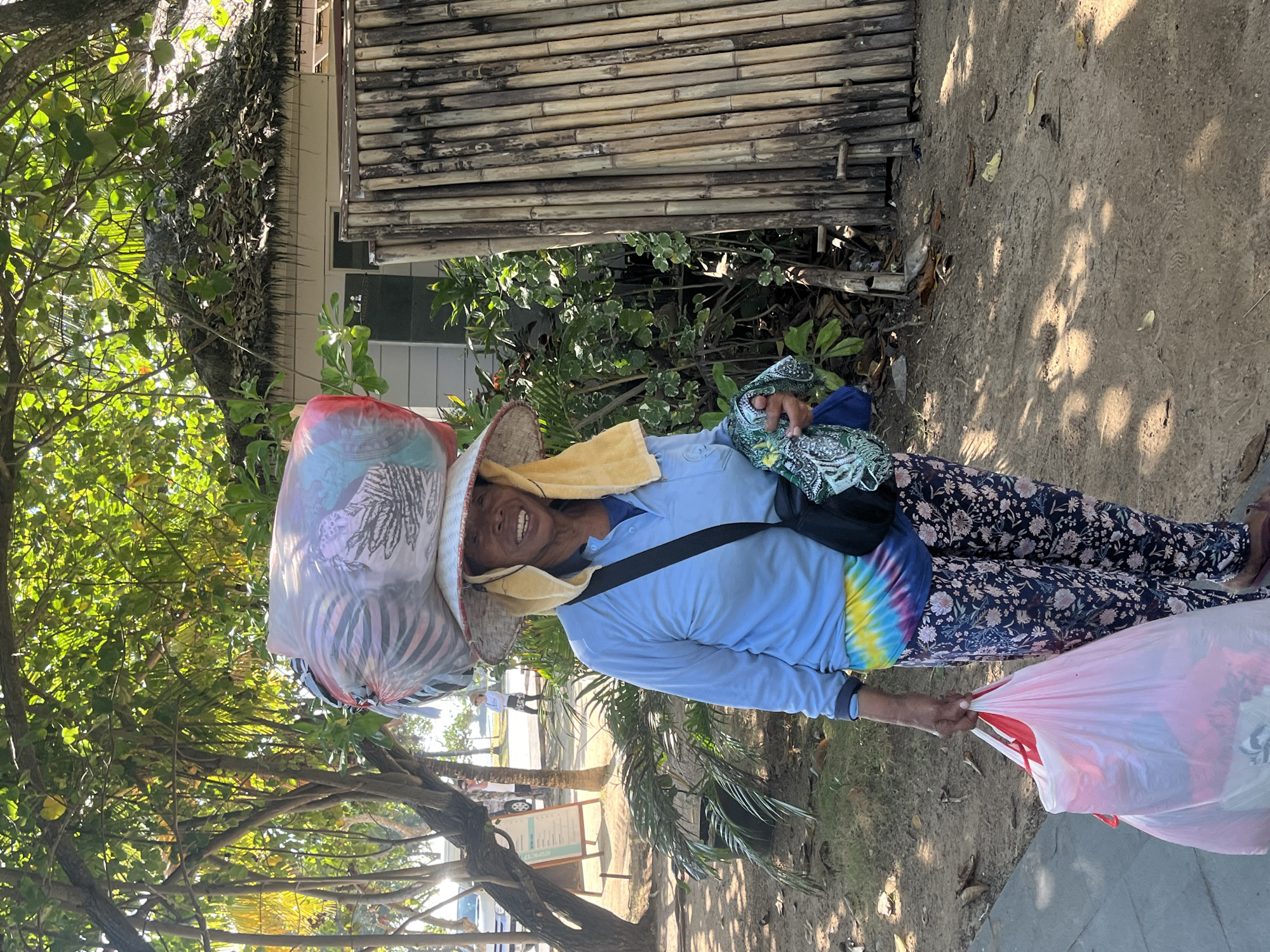 An adult female stands on a sandy path in Kuta Selatan, Indonesia, with lush green trees and a thatched-roof building in the background. She is wearing a light blue long-sleeved shirt, a tie-dye belt, and patterned pants. A large straw hat, adorned with a plastic bag filled with items, is balanced on her head. A yellow towel is draped over her shoulders, and a black bag is slung across her chest. She holds a large pink plastic bag in her right hand, and a green patterned fabric in her left. The ground is sandy and scattered with dry leaves, and sunlight casts dappled shadows. In the distance, a car and other pedestrians are visible. The overall scene suggests a busy, tropical environment.