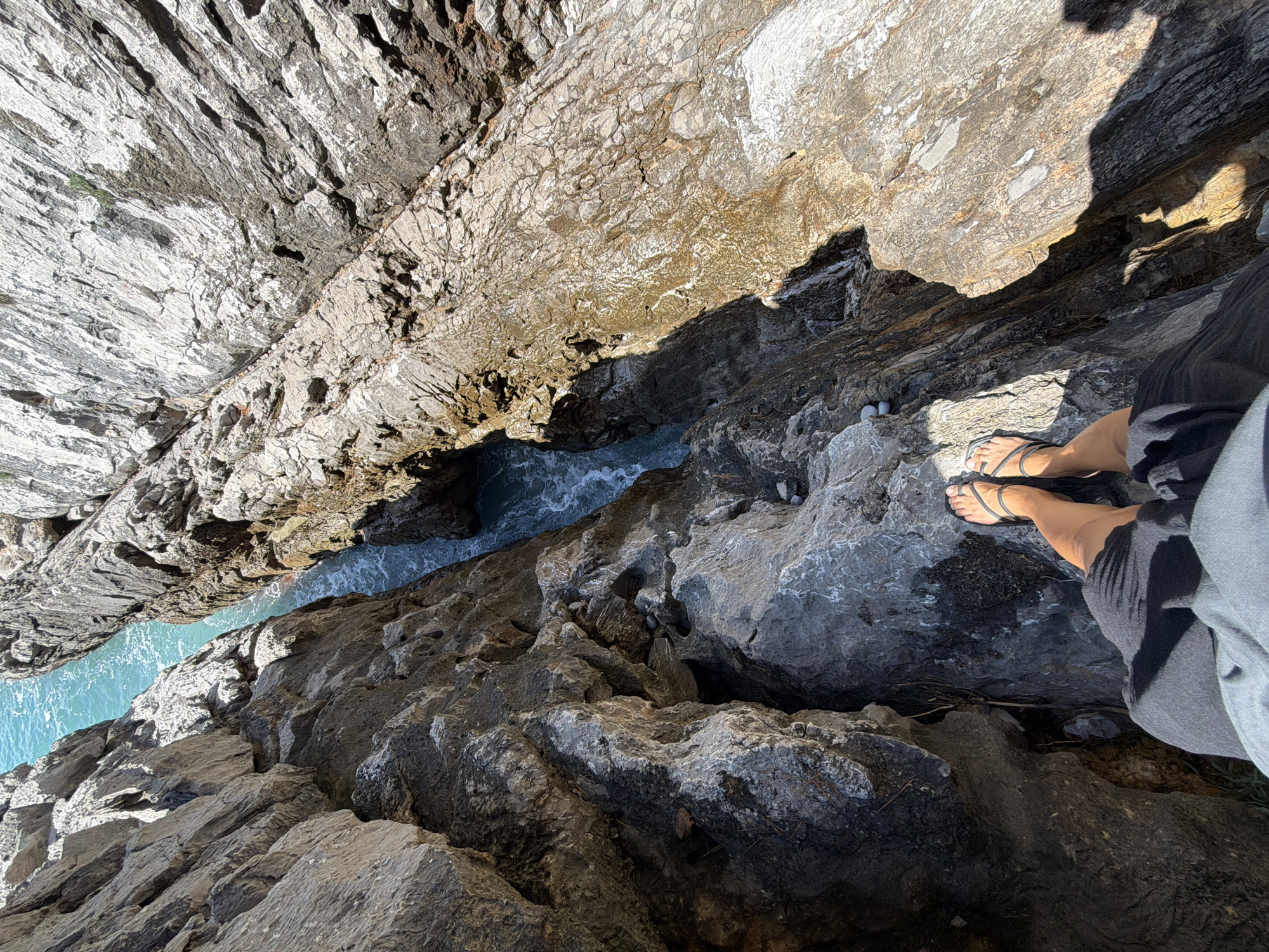 The image offers a dramatic, top-down perspective into a deep, narrow rocky gorge or sea inlet located in Boreti, Montenegro. Imposing, stratified rock formations, showcasing varying shades of grey, brown, and light beige, dominate the scene, framing a vibrant channel of turquoise water at their base. The water exhibits gentle ripples, suggesting a calm sea.
In the lower right, a person's bare legs and feet are visible, clad in black thong sandals. A dark-colored garment, possibly a skirt or shorts, partially covers their lower body. The person appears to be standing on the rocky edge, looking down into the chasm.
Bright natural sunlight illuminates the scene, casting strong, distinct shadows that accentuate the rugged textures of the rock formations, indicating a clear and sunny daytime. The overall impression is one of exploring a striking natural coastal landscape. There is no visible text in the image.