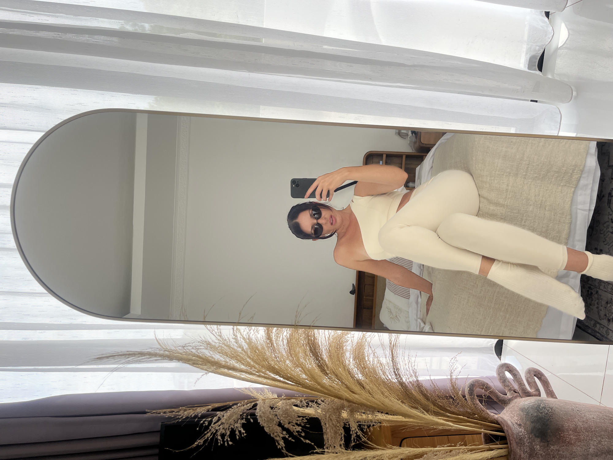 This image captures a bright, indoor mirror selfie, likely taken in a bedroom. The main subject is a person reflected in a large, arched-top, full-length mirror. The person is taking the photo with a dark smartphone, held up in one hand, and is wearing dark sunglasses. They are dressed in a cream or off-white two-piece outfit, consisting of a crop top and matching leggings, paired with light-colored socks. They are casually posed, appearing to be lying or sitting on a bed with light-colored bedding, including a textured beige blanket and white sheets. A wooden headboard is partially visible behind them in the reflection.
The room is brightly lit, suggesting it is daytime, with natural light entering through a window covered by white horizontal blinds, visible above and around the mirror. In the foreground, outside the mirror's frame, a prominent decorative arrangement of dried pampas grass or similar natural elements is displayed in a rustic, reddish-brown vase or pot. The overall aesthetic is neutral and natural, with dominant tones of white, cream, beige, and light grey. No specific emotions are discernible due to the sunglasses, and no text is visible in the image. The scene is consistent with a relaxed home environment in Kuta Selatan, Indonesia.