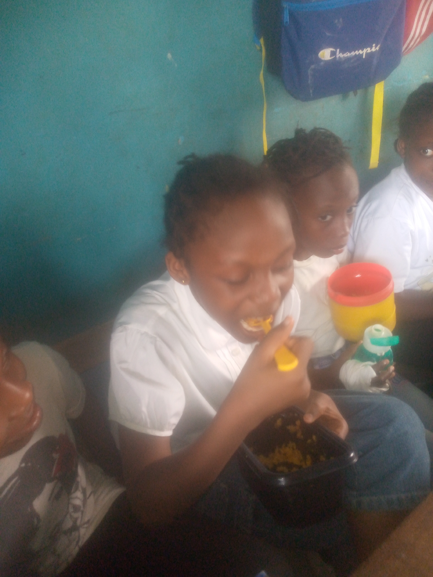 The image shows three children, presumably in a room with turquoise walls. One child is actively eating from a black container with a yellow spoon. A second child sits beside them, holding a yellow and red cup and a small toy. A third child is partially visible on the right edge of the image. All three children appear to be wearing white shirts. A blue Champion brand backpack hangs on the wall behind them. The location is described as Unknown, Unknown.