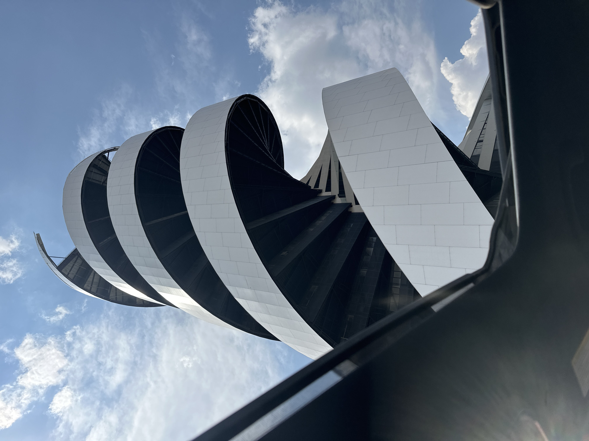 The image presents a low-angle view of a modern architectural structure, likely a building, against a backdrop of a partly cloudy blue sky. The building's design incorporates a series of curving, parallel bands or segments, alternately white and black. The white segments appear to be clad with a tile-like material, while the black segments reveal a stair-like internal structure. An out-of-focus object or structure is visible at the bottom, intruding into the view. The overall aesthetic of the building is sleek and contemporary. This is located in the city of San Bernardino Tlaxcalancingo, Mexico.