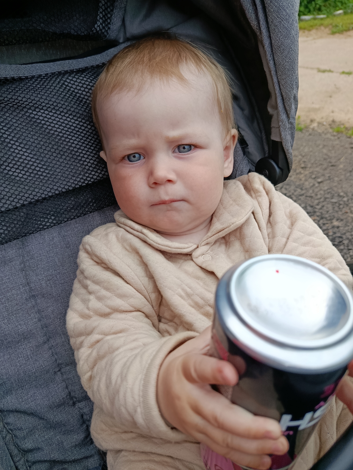 The image shows a close-up of a baby sitting in a stroller. The baby has blond hair, blue eyes, and a slightly grumpy expression. They are wearing a quilted, light brown jacket. The baby's hands are holding a can, which is partially obscured. The can is black with some pink and white lettering, and a silver top with a small red dot.
The background of the image shows the dark fabric of the stroller seat and a blurred outdoor scene with trees and a gravel path, suggesting the photo was taken during the daytime. The overall mood of the image is casual and candid, capturing a moment with the child.
There is some visible text on the can, but it is mostly unreadable due to the angle and focus. The visible letters appear to be "H" and possibly "F" or "R" followed by other characters. The context of "Фоки, Russia" indicates the general location where the photo was taken.