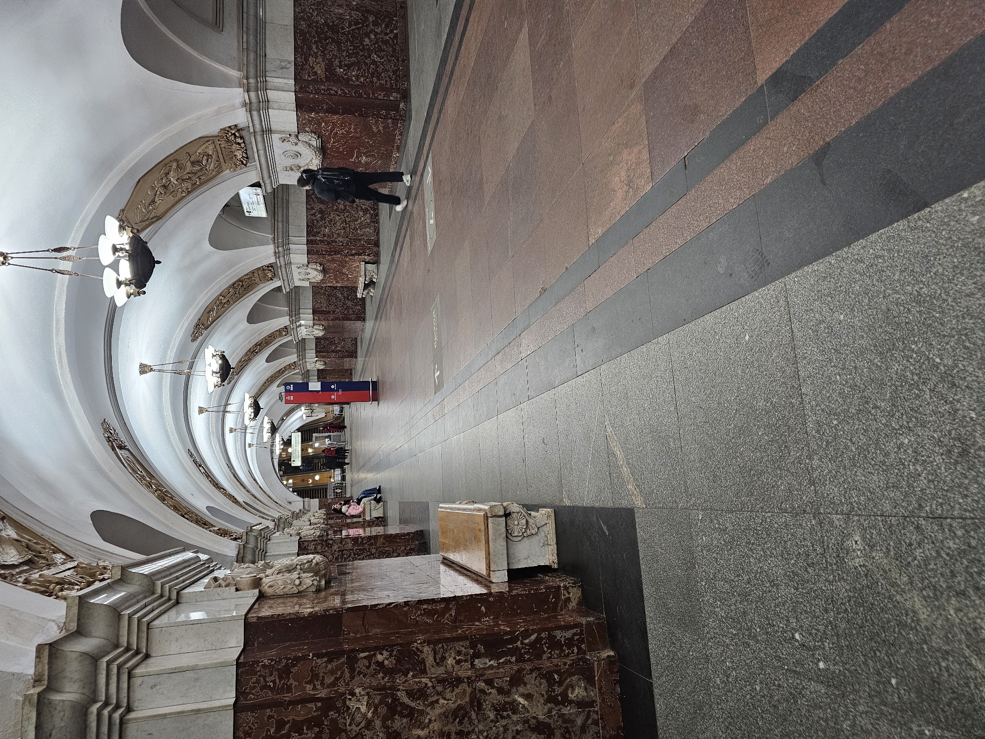 This image depicts the interior of the Ploshchad Revolyutsii (Revolution Square) Metro Station in Moscow, Russia. The scene captures a long, vaulted platform hall, illuminated by artificial light.
The architecture is characterized by a series of white, arched ceilings supported by large, ornate pillars. The pillars are clad in two distinct marble types: white or light grey marble on the upper sections and a dark reddish-brown, possibly veined marble on the lower portions. Between the arches, above the pillars, elaborate gold-colored bas-reliefs depicting figures and scenes are set into the spandrels. Multi-armed bronze chandeliers with frosted glass shades hang centrally from each arch, providing illumination.
The floor is tiled with large rectangular stone slabs, forming a pattern of dark grey, lighter grey, and reddish-brown sections. A prominent black strip runs longitudinally down the center of the visible platform area.
Several individuals are present. In the mid-right, a single individual is walking away from the camera, dressed in dark clothing (jacket, trousers) and white sneakers, carrying a black backpack. On the left side of the platform, two individuals – an adult in a light pink top and blue jeans, and a child in a pink top and dark trousers – are seated on an ornate white marble bench with a wooden top. Further down the hall, towards the background, indistinct figures are visible near a set of ascending escalators.
Additional objects include multiple similar white marble benches with wooden tops positioned along the left wall. A vertical red and blue information or directional pillar stands in the mid-right section of the platform. A smaller white rectangular sign with text is visible hanging above the escalators in the far background. Floor markings, including arrows and text, are discernible on the tiles.