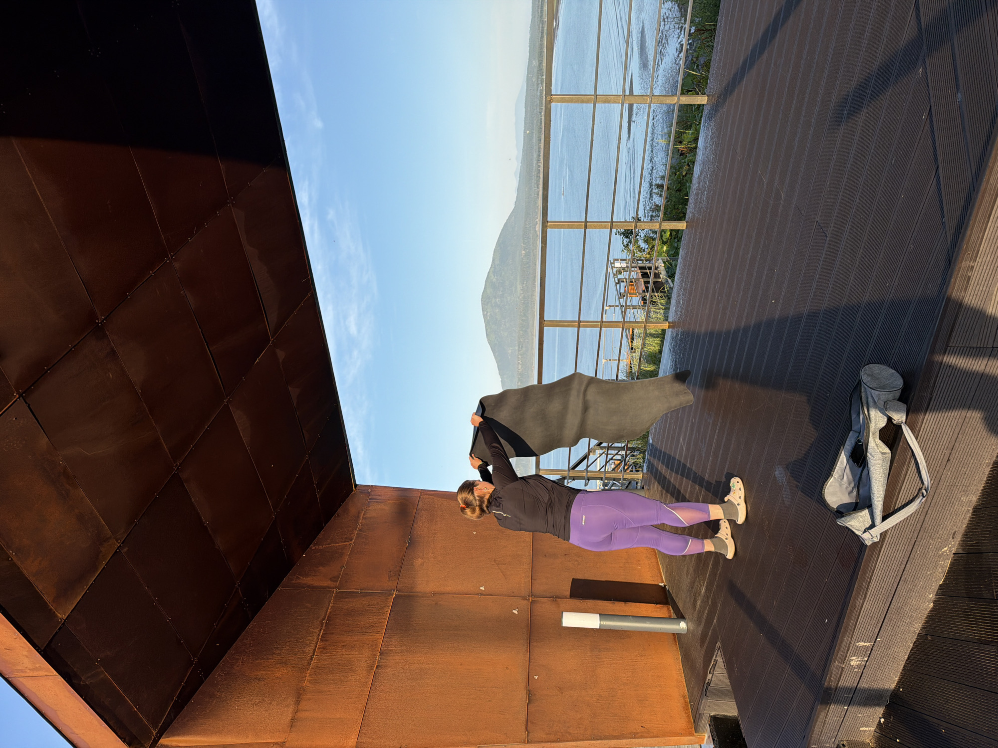 An adult woman with blonde hair tied back is standing on a dark wooden deck, holding a dark yoga mat. She is wearing purple leggings and a black long-sleeved shirt. To her left, there is a silver cylindrical light fixture. The deck is part of an outdoor structure with rusted metal walls and ceiling, offering a view of a body of water and a distant, green, conical mountain under a clear blue sky. A gray yoga mat bag is on the deck in the foreground. This scene is located in Yuzhno-Kurilsk, Russia.