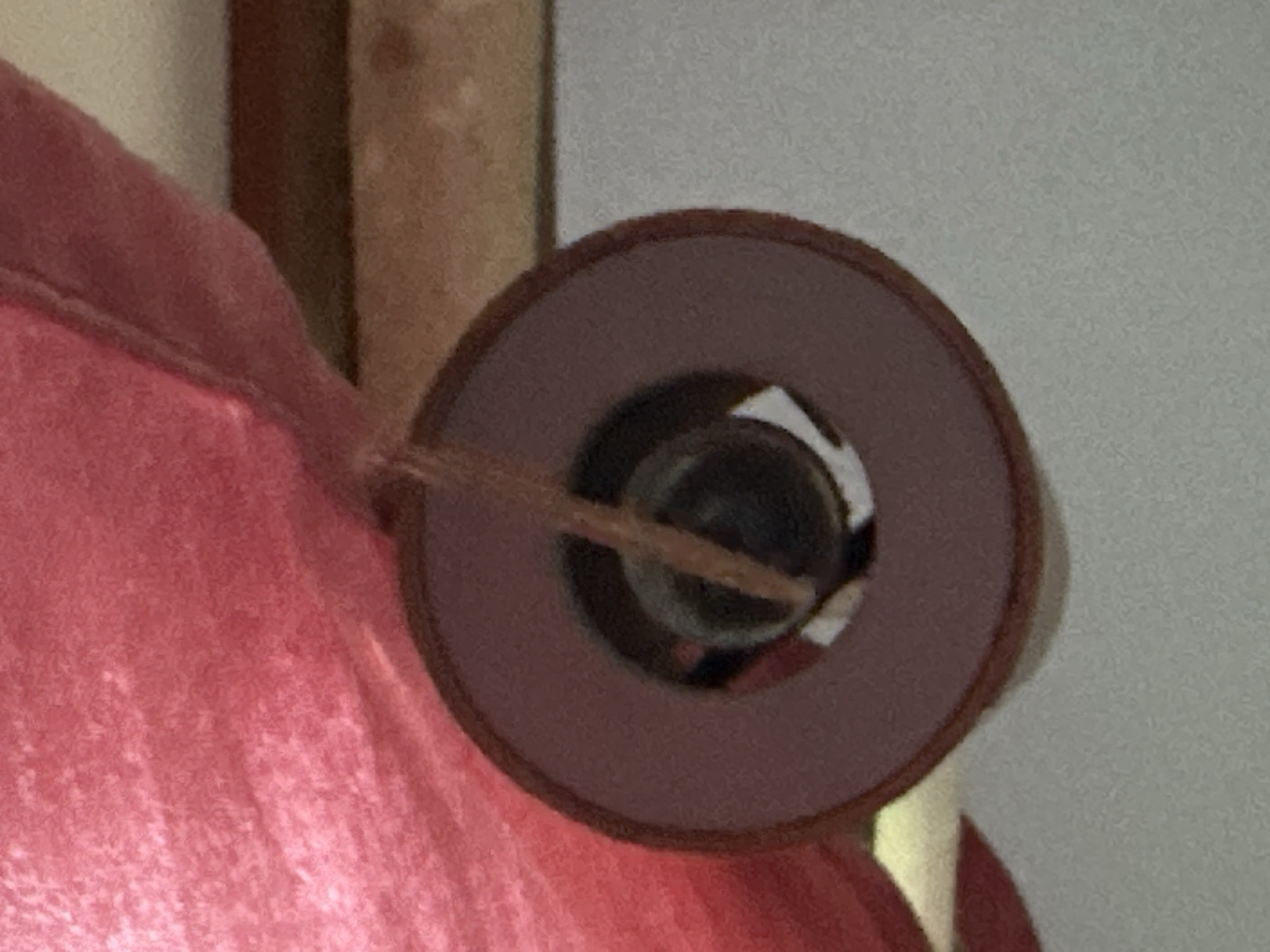 The image shows a close-up, slightly out-of-focus view of a wind chime. The wind chime has a circular, maroon-colored frame with a hollow center. Inside the hollow center, a dark, possibly metallic, object is visible. A thin, light-brown string or cord hangs from the top, passing through the center and appearing to be attached to the inner object. A blurry, pink, textured material, possibly fabric, is visible in the upper left portion of the image, suggesting the wind chime is hanging from something. The background consists of a textured surface in shades of brown and gray, which could be a wall or ceiling. There is no visible indication of the city of Thora, Australia, in this image.