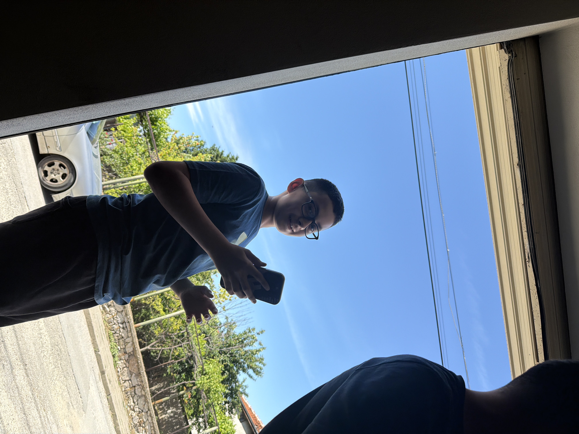 A young person with glasses is looking at a phone in an outdoor setting. The person is wearing a blue t-shirt and dark pants. A car and a stone wall can be seen in the background. The sky is blue with electrical wires visible. A second person is partially visible in the bottom of the frame. The location is presumed to be Asenovgrad, Bulgaria, based on the prompt.