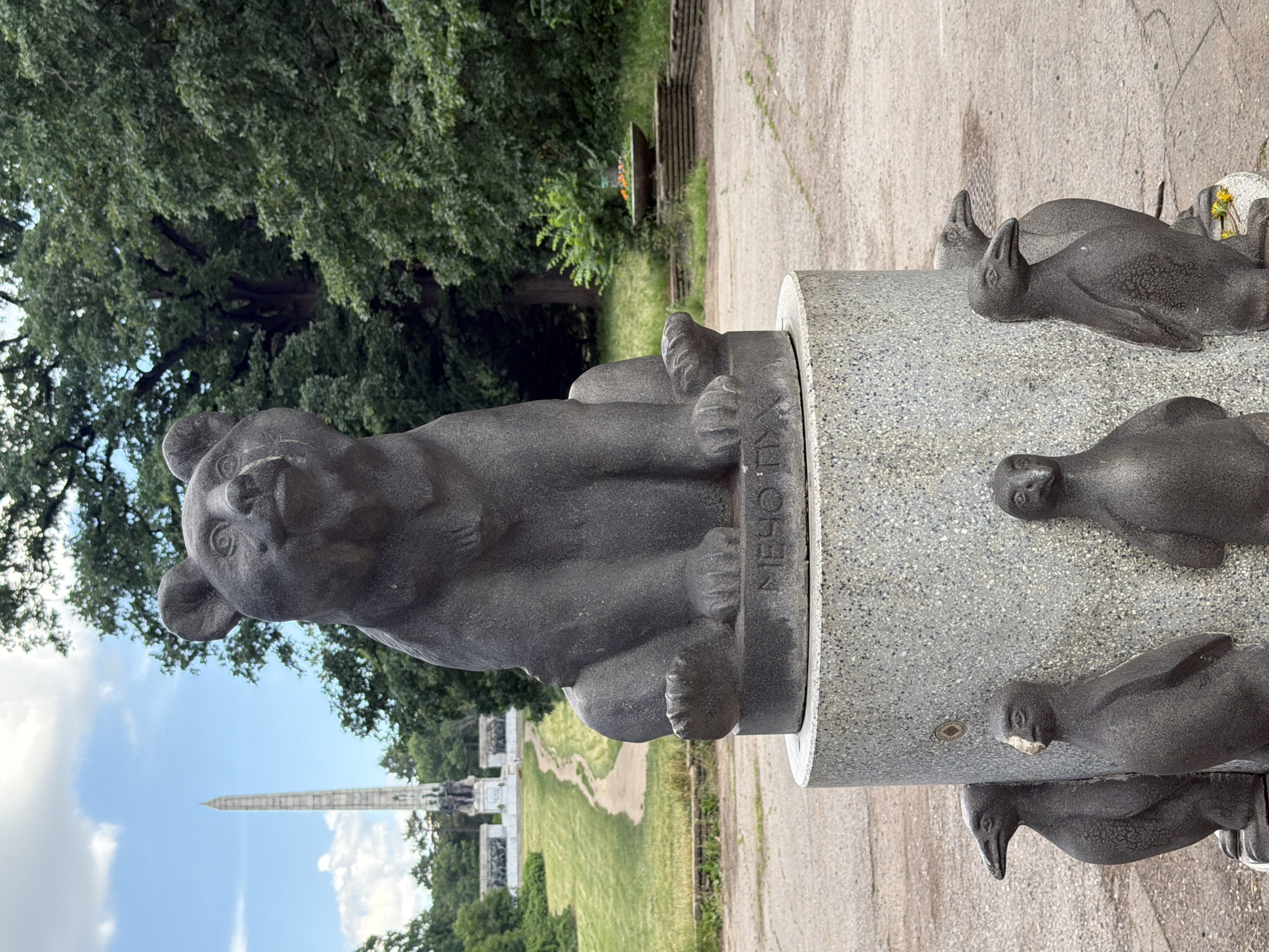 The media file features a statue of a bear, likely made of dark stone, in an outdoor setting. The bear is depicted sitting, with its paws resting on a cylindrical stone base. The base is engraved with text that reads "МЕЧО ТИ". Around the base, there are several smaller, dark stone sculptures of birds, possibly penguins or seals.
In the background, a tall, slender obelisk-like monument is visible against a bright blue sky with scattered white clouds. Lush green trees and grass surround the area, suggesting a park or garden. A pathway is visible winding through the green space. The scene appears to be set during the daytime, with bright lighting indicating fair weather.
The overall impression is of a memorial or decorative sculpture within a public space. The bear statue's expression is somewhat detailed, giving it a mild or neutral appearance. The smaller animal sculptures add a whimsical or thematic element to the monument.