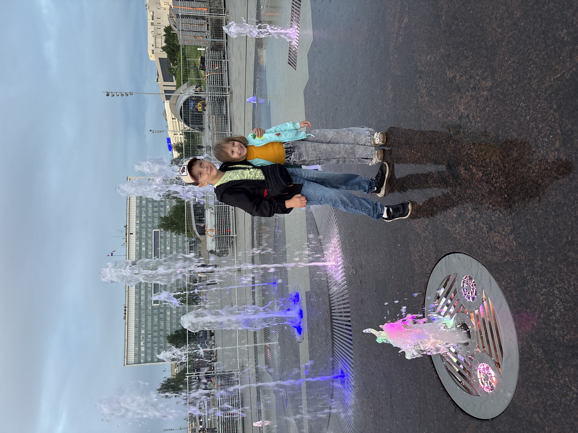 This image captures a wide shot of two children posing in front of a series of illuminated interactive fountains within a public square. A boy, positioned on the left, wears a black jacket over a light green t-shirt, blue jeans, and black sneakers, topped with a dark baseball cap featuring a light emblem; a black fanny pack crosses his chest. To his right, a younger girl wears a yellow ribbed long-sleeve top, a light blue jacket with a small pineapple motif, light grey wide-leg jeans, and light-colored slip-on shoes. Both children are smiling broadly at the camera, with the girl's right arm gently around the boy's lower back.
The ground beneath them is a dark, highly reflective, wet stone surface, showcasing clear reflections of the children and the surrounding lights. Numerous fountains are actively spraying water vertically from in-ground fixtures. Many of these fountains are dynamically illuminated with colored lights, displaying hues of purple, pink, blue, green, and white. A prominent fountain in the lower-left foreground emits a vivid, multi-colored spray.
In the mid-ground, a line of white temporary metal barriers or fencing separates the immediate fountain area from what appears to be an event setup. Behind these barriers, stacks of event infrastructure, possibly stage elements or seating, are visible, along with some yellow machinery and a large, curved, tent-like structure.
The background features several urban buildings beneath an overcast sky. A large, multi-story rectangular building with a grey facade and numerous windows is prominent on the left, with two flags visible on its roof, one of which appears to be the Russian national flag. To the right, another lighter-colored, modern building is visible, alongside tall light poles.
The scene is located at the **Perm Esplanade** in Perm, Russia. The distinctive large grey building in the background is the **Government of Perm Krai building** (ул. Ленина, 51).