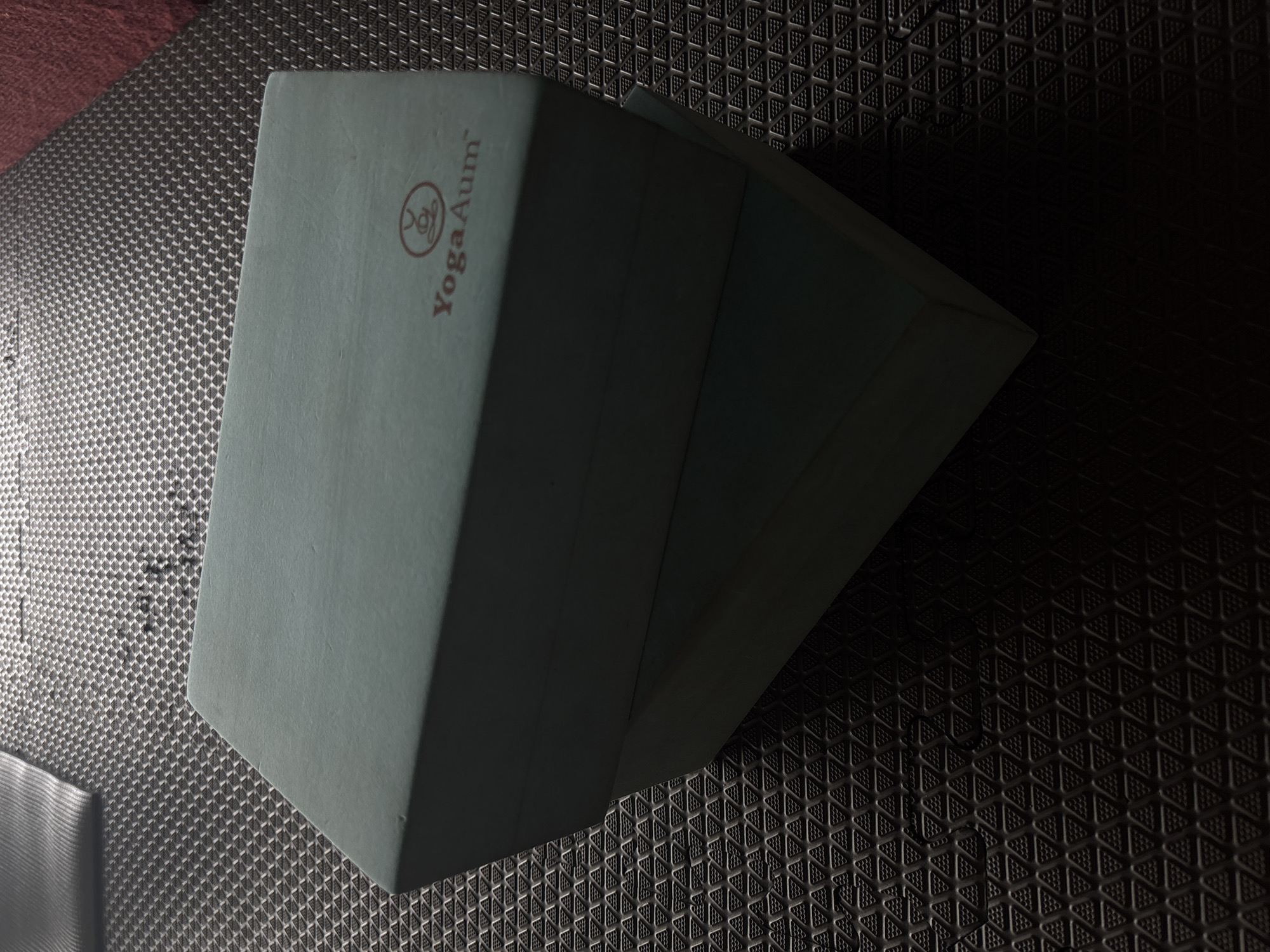 The image shows two light blue yoga blocks stacked on a dark gray yoga mat with a triangular pattern. The block on top has the "YogaAum" logo in brown. The mat is likely in a studio or home setting. The location in Rawai, Thailand is not evident from the image alone.