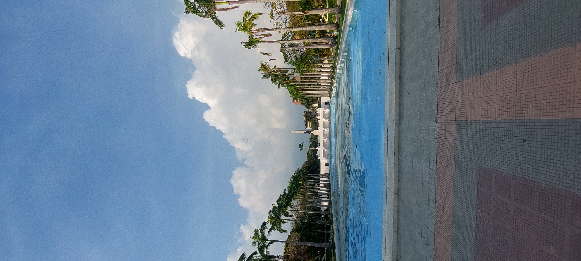 The image captures a grand outdoor monumental landscape under a bright, partly cloudy sky. A long, narrow, rectangular pool or water feature with a light blue bottom occupies the midground, its surface reflecting the clear blue sky and some white cumulus clouds.
On the left side of the water feature, a neat row of tall palm trees with green fronds extends into the distance, lining the path alongside the pool. In the background, beyond the palm trees and pool, several white monumental structures are visible, including a prominent obelisk and a dark-colored statue, possibly equestrian, situated in front of it. Further white buildings with columns can also be seen.
To the right of the water feature, a textured pathway or ground area is visible, paved with rectangular tiles in shades of gray and reddish-brown, arranged in a grid-like pattern.
The scene suggests a sunny daytime setting with fair weather. No individuals or specific activities are present, lending a sense of stillness and grandeur to the environment. There is no visible text. The overall composition points to a significant public space, characteristic of monumental areas in Caracas, Venezuela, such as the Paseo Los Próceres.