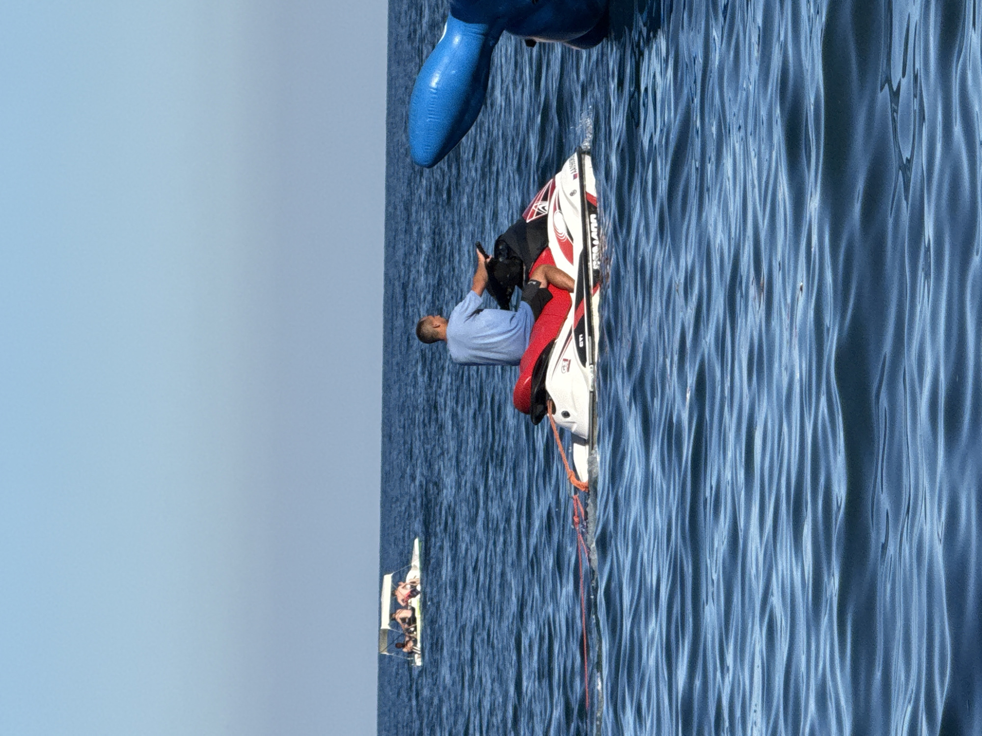 The image depicts a body of water, likely the sea, under a clear blue sky. In the foreground, a man is riding a red and white jet ski, with a rope attached to its rear, pulling something unseen. To the left, further in the distance, a small white boat with an awning is visible, carrying at least three people. To the right of the jet ski, a large blue inflatable object, possibly part of a water attraction, is partially visible. The water is blue and shows gentle waves. The scene appears to be a recreational area on a sunny day. The image is taken in the city of Partenit, Ukraine.