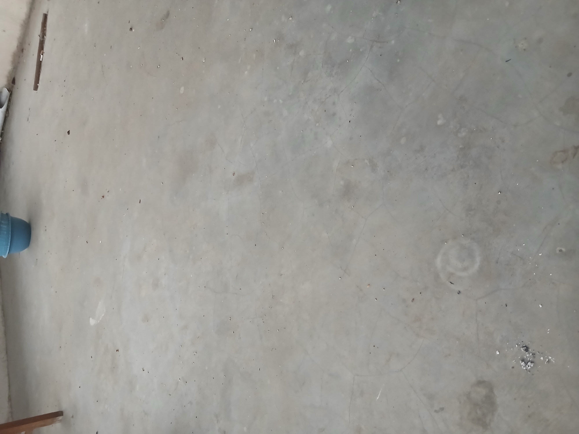 The image depicts an outdoor concrete floor, possibly a patio or walkway, in Tenggarang, Indonesia. The surface is predominantly gray with visible cracks and some water stains. A blue plastic bucket is situated in the upper right corner of the frame. To the left, a portion of a wooden table or chair leg is visible. Scattered debris, including small stones and what appears to be ash or dust, is present across the floor. The lighting suggests it is daytime. There are no people or animals visible in the scene.