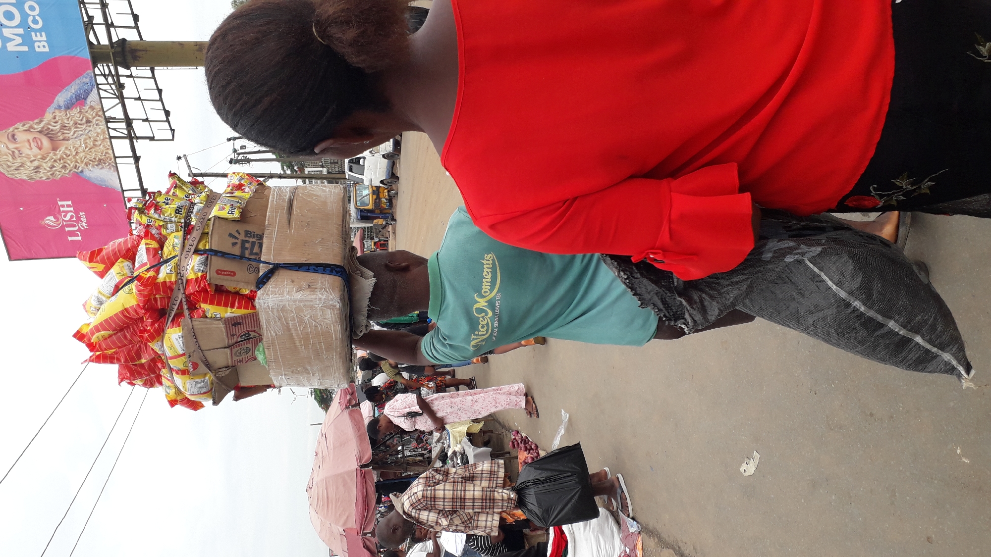 An eye-level outdoor shot captures the bustling commerce of Onitsha, Nigeria, with a focus on a man carrying a load of goods on his head. The man, clad in a green T-shirt with the words "Nice Moments" and the phrase "NATURAL SENNA LEAVES TEA," balances wrapped boxes and bags of what appear to be snacks or food items. A woman with black hair styled in a bun stands to his side wearing a red blouse.
The ground is gray and appears to be pavement with vendors, merchants, and shoppers in the background. Some people are seated while others stand. Displayed on tables are a variety of items.
A large advertisement for "LUSH Hair" looms in the background. The sky is an overcast white.