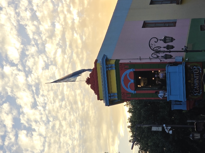 A vibrant building in La Boca, Argentina, stands tall against a sky filled with clouds at sunset. The building features colorful paint, intricate architectural details, and the Argentine flag waving from its rooftop. A lamp post and a sign reading "Caminito" are visible. People gather inside, visible through a window. The surrounding scenery includes greenery and the facades of other buildings, creating a lively and picturesque scene.