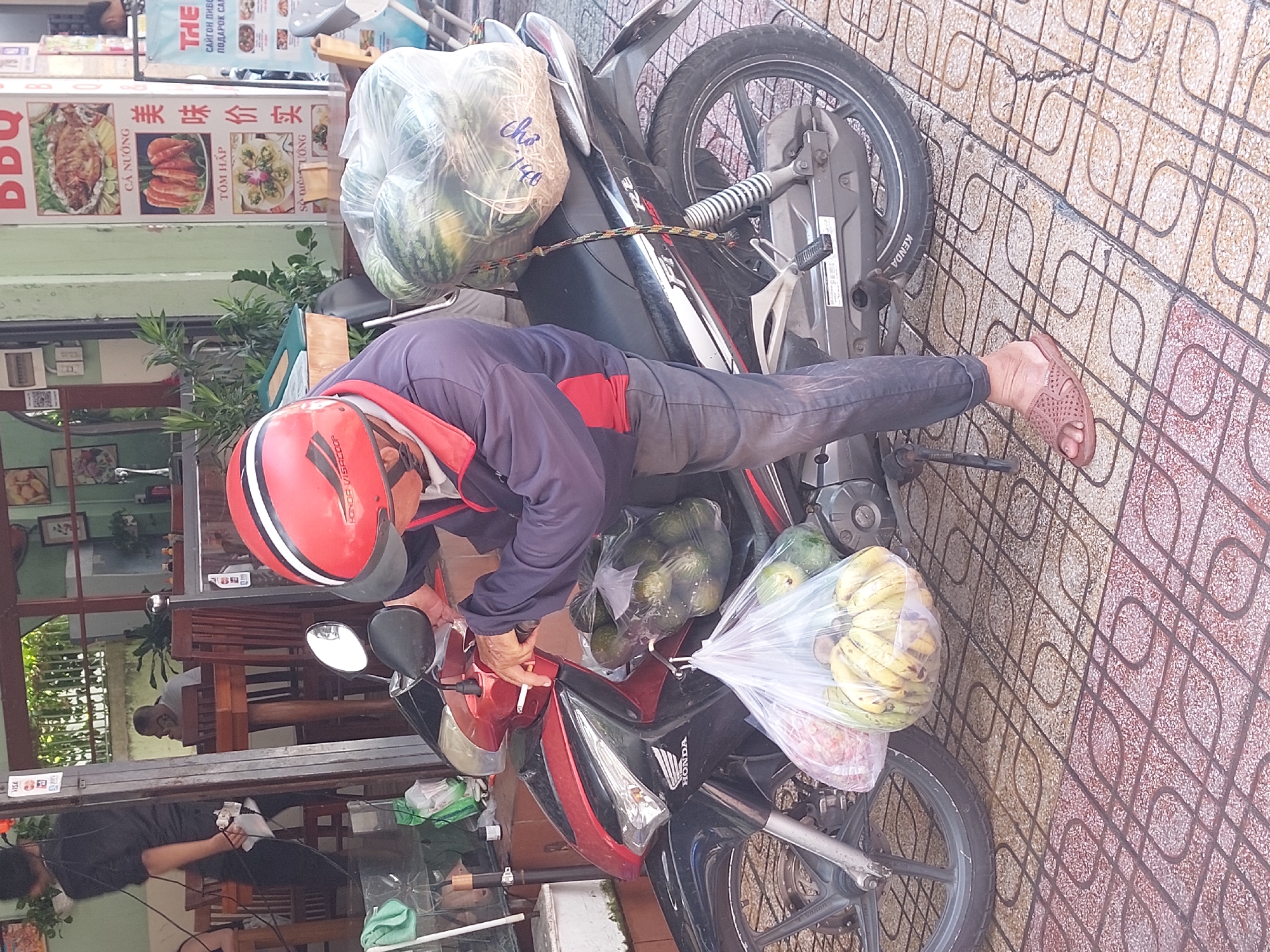 A man is standing next to a red and black motorcycle in what appears to be a street in Nha Trang, Vietnam. He is wearing a red and white helmet, a dark blue jacket with red accents, and dark gray pants. He is holding onto the handlebars of the motorcycle and has a cigarette in his right hand. A large bag of watermelons is tied to the back of the motorcycle, and two plastic bags filled with fruit, possibly bananas and possibly some kind of red fruit, are hanging from the side of the motorcycle. The man is wearing brown sandals. The motorcycle is parked on a tiled sidewalk, and a portion of a building with signage can be seen in the background. The overall scene suggests a moment of pause or preparation for transport of goods.