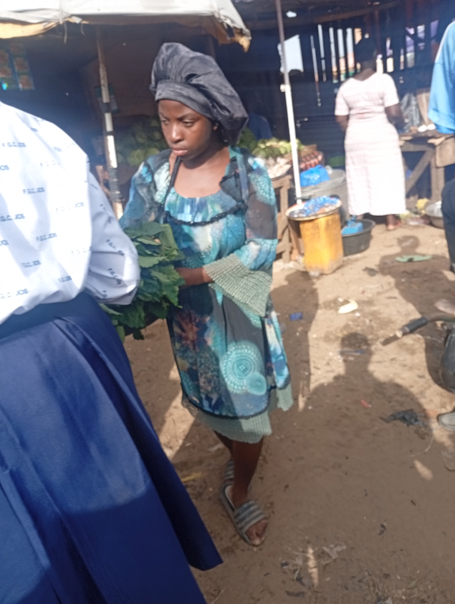 This image depicts a scene in an outdoor market, likely in Rafin Par, Nigeria. A young woman wearing a dark head covering and a blue and teal patterned dress is the central figure. She is holding a bundle of leafy green vegetables in her arms. To her left, the arm and shoulder of another person are visible, wearing a white shirt with blue lettering. The ground is sandy and appears well-trodden. In the background, there are market stalls with wooden structures and awnings, suggesting a bustling commercial area. Other individuals are partially visible, including a woman in a light-colored dress standing further back and the legs of another person in blue. The overall environment suggests a typical market day in a Nigerian town.