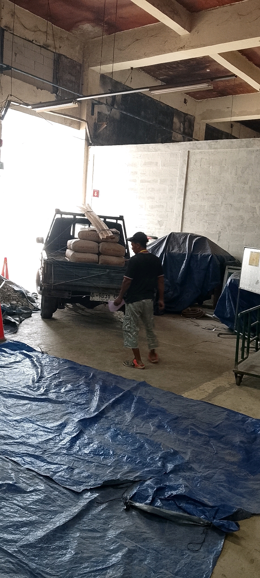 In Depok, Indonesia, a man wearing a black t-shirt and camouflage shorts, with a black baseball cap, walks away from a black pickup truck loaded with sacks of cement and wooden planks. The truck is parked indoors, likely in a warehouse or construction site, as evidenced by the concrete floor and unfinished walls. In the foreground, a large blue tarp is spread out on the ground, partially obscuring the truck's rear wheels and the surrounding area. Other construction materials and debris are visible, including a pile of gravel to the left and a tarp-covered object to the right. The lighting suggests an overcast day or artificial lighting within the structure.