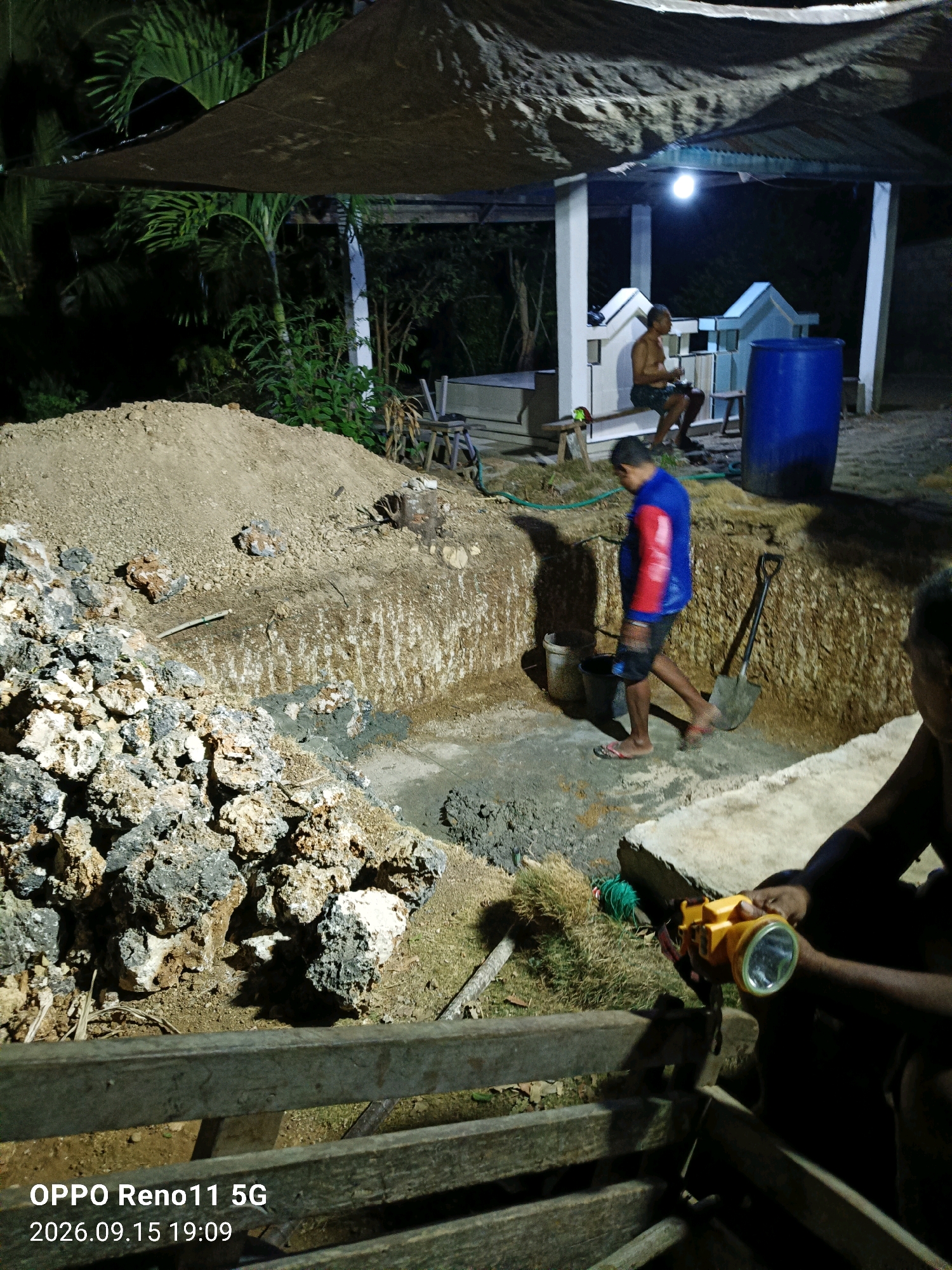 The image depicts an outdoor scene at night, likely in a rural or undeveloped area, possibly Otan, Indonesia, given the timestamp. The primary activity appears to be excavation, with a large, deep pit dug into the earth. Piles of soil and rocks surround the excavation site. In the foreground, a person is holding a yellow flashlight.
Further into the scene, a man in a blue and red jacket is walking near the pit, and a shovel is leaning against the wall of the excavation. In the background, a covered structure with white pillars is visible, housing another individual sitting on a bench and a large blue barrel. Palm trees and dense vegetation are present, suggesting a tropical environment. The lighting is dim, with artificial lights from the structure and the flashlight providing illumination. The timestamp on the image indicates it was taken on September 15, 2026, at 19:09.