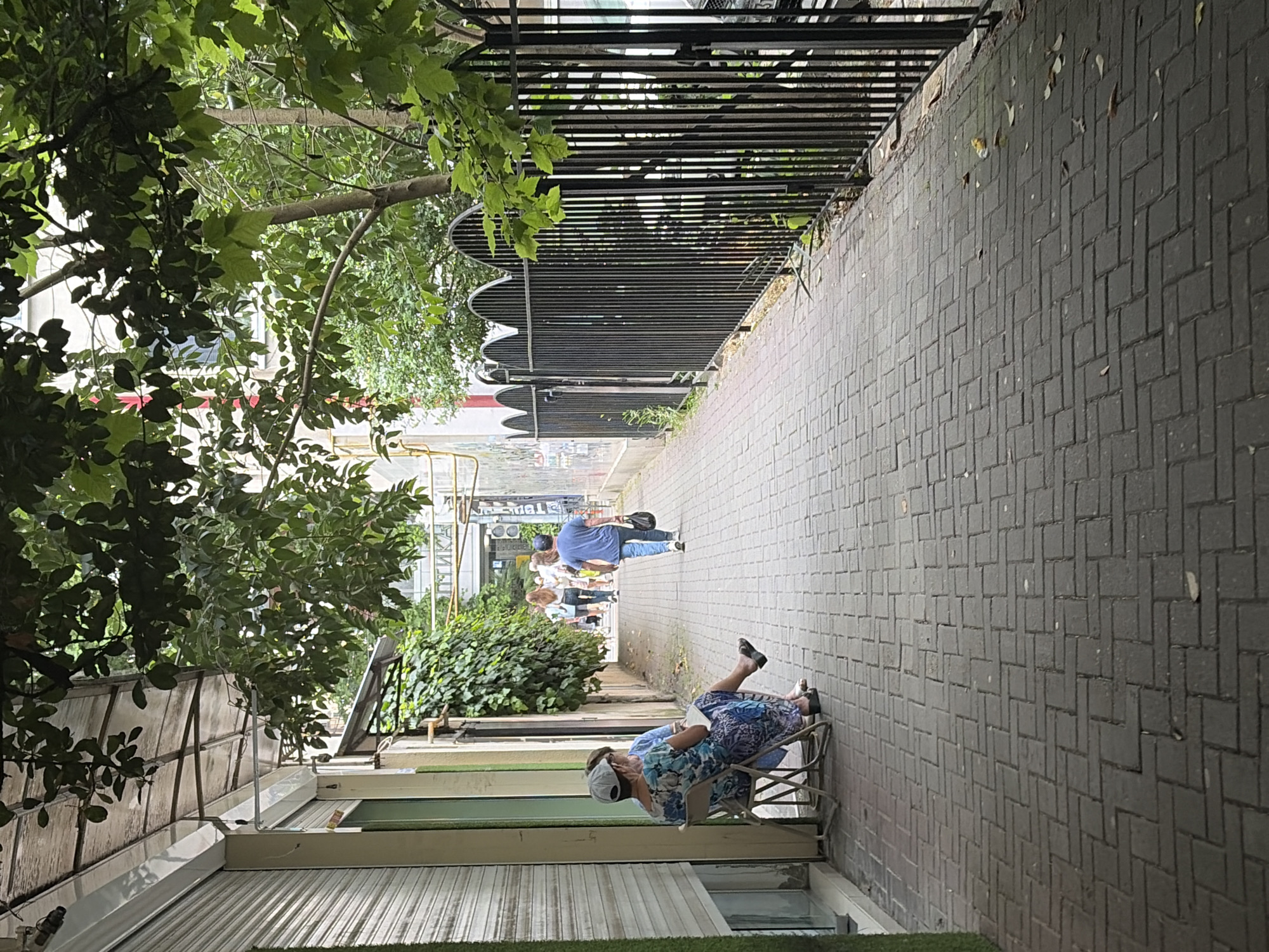 The image depicts a sidewalk scene in Sochi, Russia. The perspective is tilted, giving a sense of looking down a pathway. On the right, a grey brick-paved sidewalk stretches into the distance. On the left, lush green foliage from trees and bushes lines the path. Beyond the foliage, buildings with large windows and light-colored facades are visible, along with some industrial elements like corrugated metal roofing. A black metal fence with horizontal bars runs along the edge of the sidewalk.
Two people are present. In the foreground, a person wearing a floral-patterned dress and a white cap sits on a metal chair, appearing to read a book. Further down the sidewalk, another person in a blue t-shirt and jeans walks away from the camera, carrying a black bag. A group of people are also visible in the background, walking along the sidewalk.
The scene appears to be set during the daytime, with overcast or diffused lighting. There are no clear indicators of the time of day or specific weather conditions, other than the general brightness suggesting it's not night. The presence of trees and greenery suggests a mild climate, consistent with Sochi. No text is clearly legible in the image.