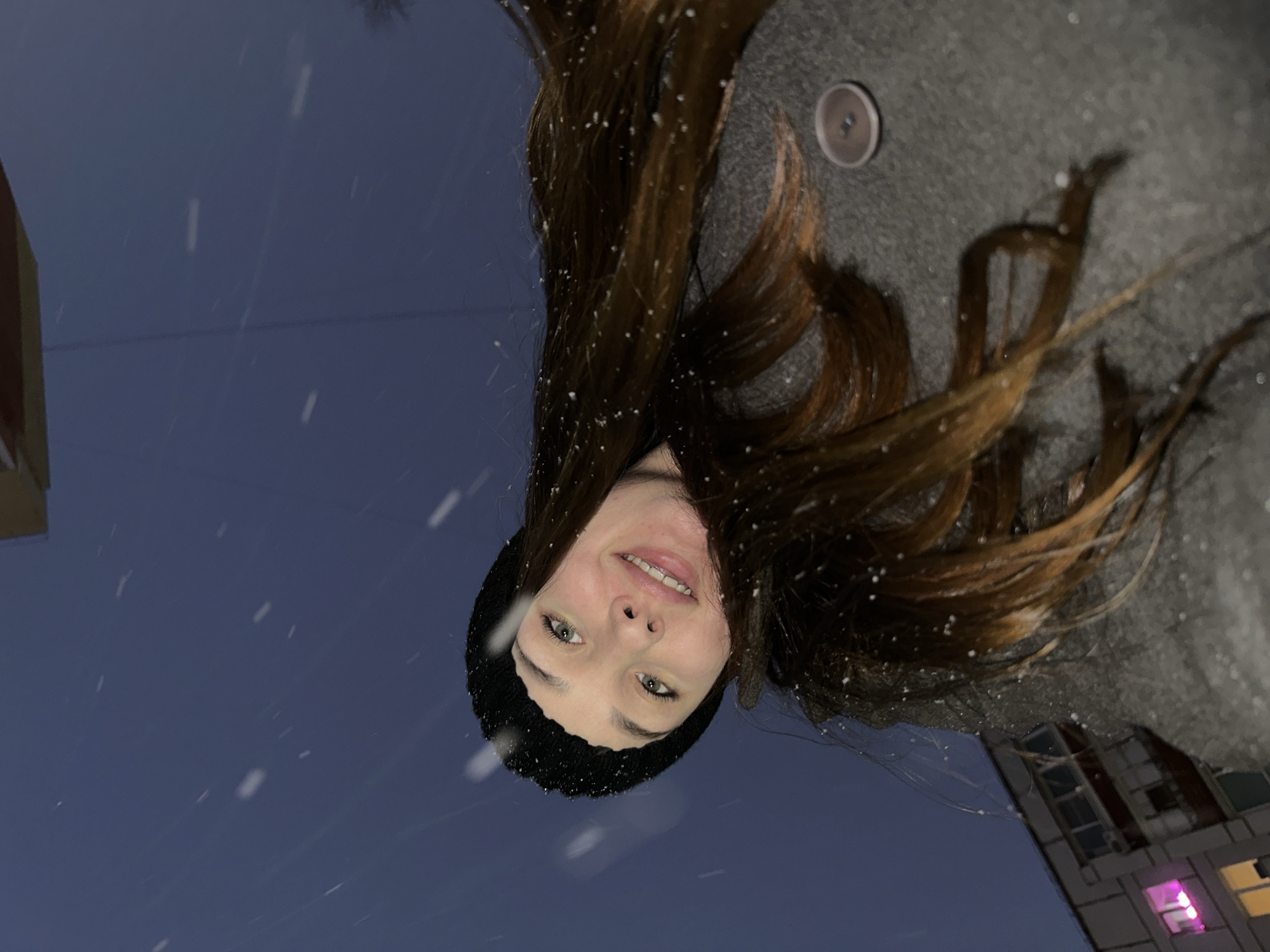 A low-angle, outdoor photograph shows a young woman with brown hair wearing a black beanie, a gray coat, and smiling slightly. It appears to be snowing. In the background, there's a building. Based on the architectural style and apparent lighting, this scene may be from the city of Chaikovsky, Russia.
