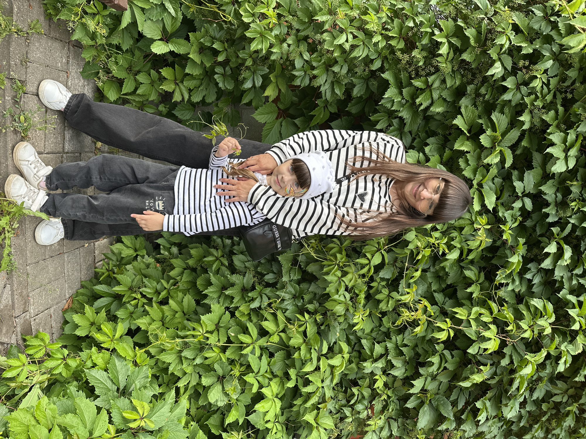 A woman and a young child stand close together in front of a dense wall of green ivy. The woman, with long brown hair and bangs, wears glasses and a black and white striped long-sleeved shirt. She has her right arm around the child. The child, wearing a white bandana, a black and white striped long-sleeved shirt, and dark pants, looks towards the camera with face paint on their cheek. Both individuals are wearing white sneakers. The child is holding a small green leafy branch in their right hand. A black crossbody bag with the word "GUESS" is visible on the woman's left side. The ground beneath them is paved with gray stones, and some small weeds are growing between them. The ivy wall forms a verdant backdrop, suggesting an outdoor setting. The image was taken in Izhevsk, Russia.