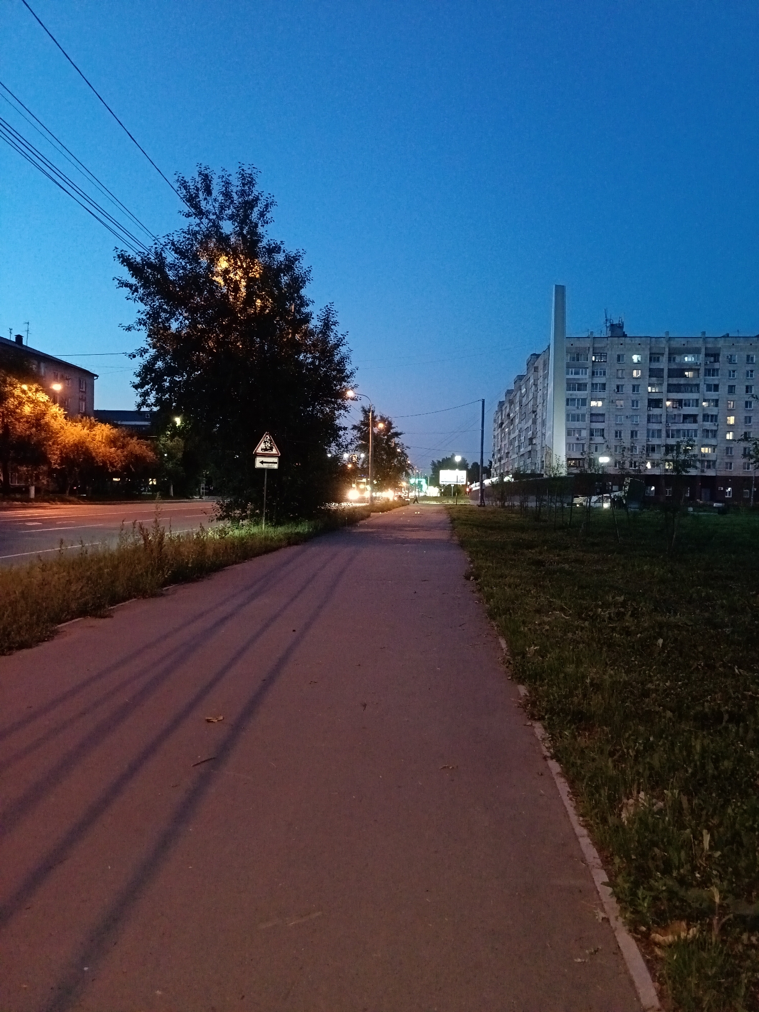 An urban scene at dusk or dawn is depicted, likely in Komsomolsk-on-Amur, Russia. The sky is a deep blue, transitioning from lighter shades near the horizon. A paved sidewalk runs along the right side of the image, bordered by a grassy area with some small plants. To the left of the sidewalk is a paved road with visible lane markings.
Along the road, a dense tree with illuminated foliage stands prominently. Across the road, a large, multi-story apartment building dominates the background. It is a long, block-style structure with numerous windows, many of which are lit, suggesting occupancy. To the left of the apartment building, another building is partially visible, with streetlights illuminating its facade.
Several streetlights are present throughout the scene, casting pools of light on the road and sidewalk, and creating long shadows from the trees on the pavement. A traffic sign with a yellow triangle and a depiction of a vehicle is visible near the large tree. In the distance, headlights from vehicles on the road indicate ongoing traffic. Power lines are also visible stretching across the sky. The overall impression is of a residential area at a transitional time of day, with artificial lighting beginning to dominate the natural light.