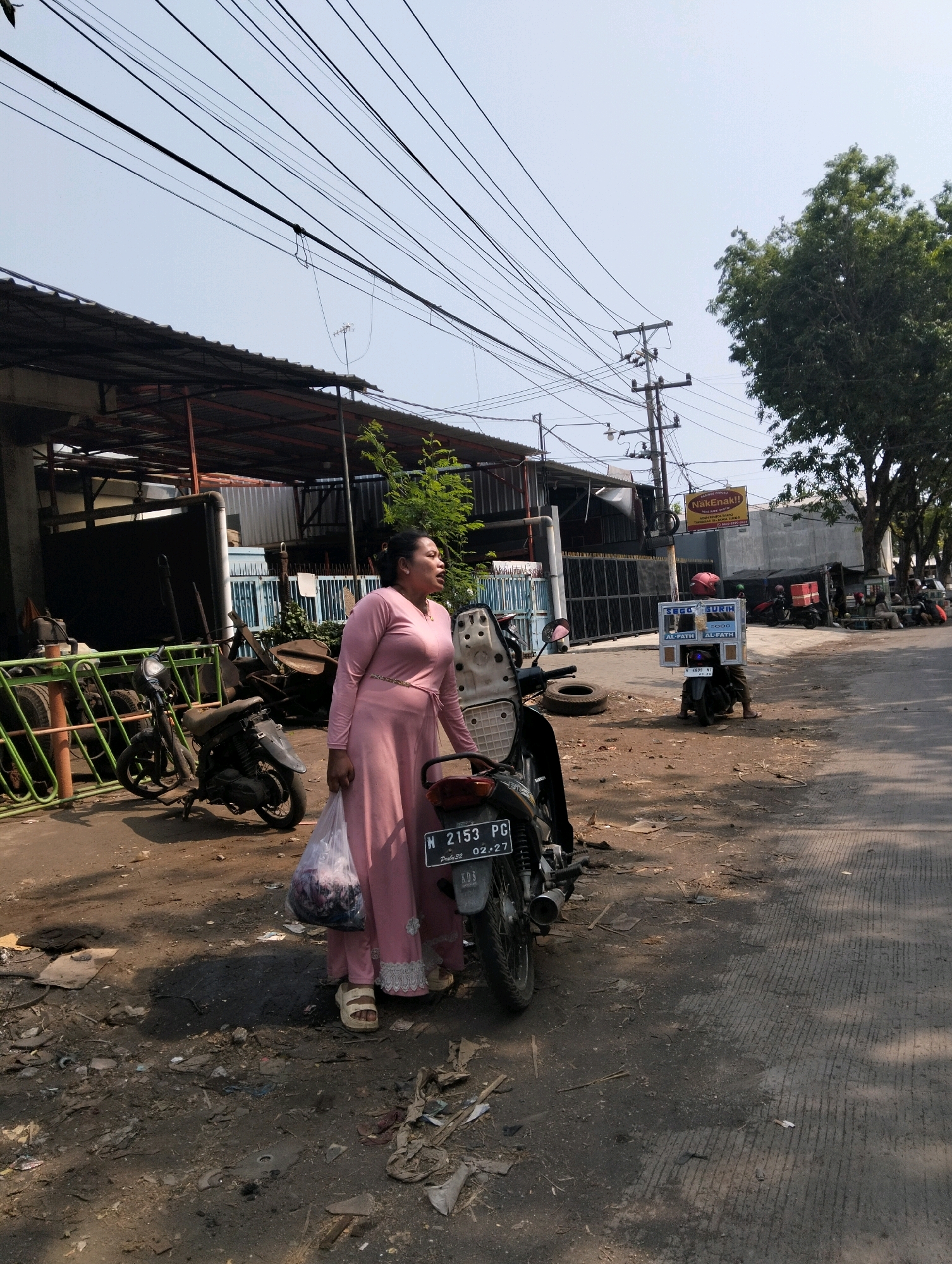 An adult female in a light pink dress stands next to a parked motorcycle on a dirt and asphalt road in Probolinggo, Indonesia. The woman is holding a clear plastic bag filled with items. The motorcycle, a black Honda with a license plate reading "N 2153 PG 02-27", is parked in front of her. In the background, there is a building with a corrugated metal roof, several other parked motorcycles, and a utility pole with numerous overhead wires. A tree with green foliage is visible on the right side of the image. The sky is clear and bright.