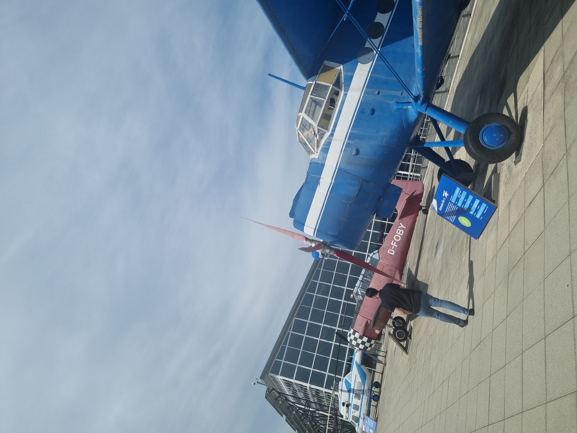The image depicts two aircraft on display outside the Flugwelt Stuttgart aviation museum in Echterdingen, Germany. The primary aircraft is a blue and white Antonov An-2 biplane. Beneath it, a red and white aircraft is partially visible, with the registration "D-FOBY" painted on its fuselage. A person wearing a black shirt and jeans stands near the red and white aircraft, seemingly observing it. In the background, a modern glass and steel building is visible. The ground is paved with light-colored rectangular tiles. The sky is light blue with some clouds. A blue and white helicopter is visible in the distance. A blue sign is near the aircraft.