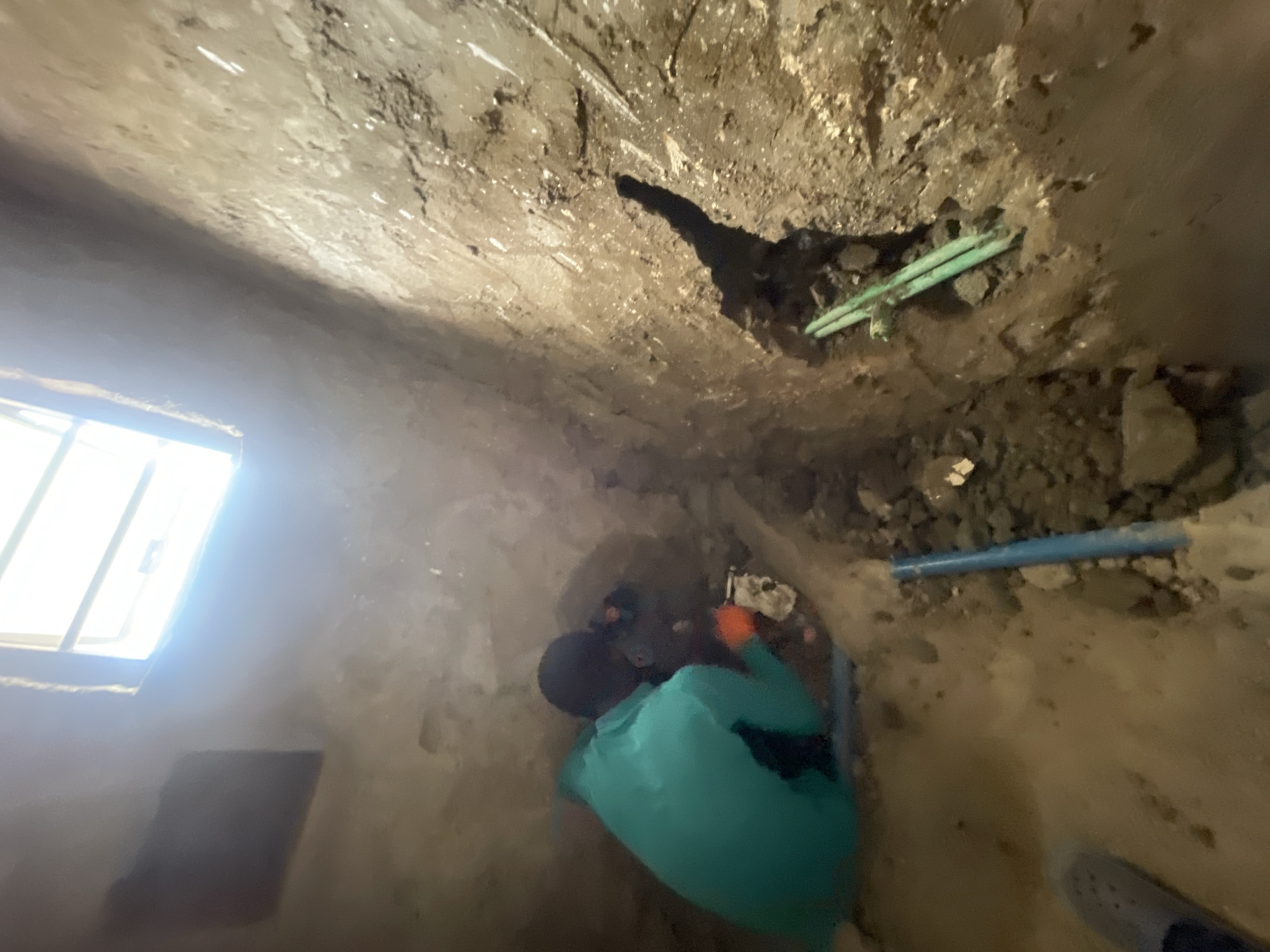 The image depicts an indoor plumbing repair situation, potentially in Angwan Sayawa, Nigeria. A person wearing a teal shirt and gloves is working on exposed pipes. The environment consists of what appears to be unfinished walls with visible dirt, pipes, and a window on the left. There are two green pipes near the top right of the image. There is also a single blue pipe near the center right of the image. The person appears to be making a plumbing connection in what could be a crawl space or service area under the floor.