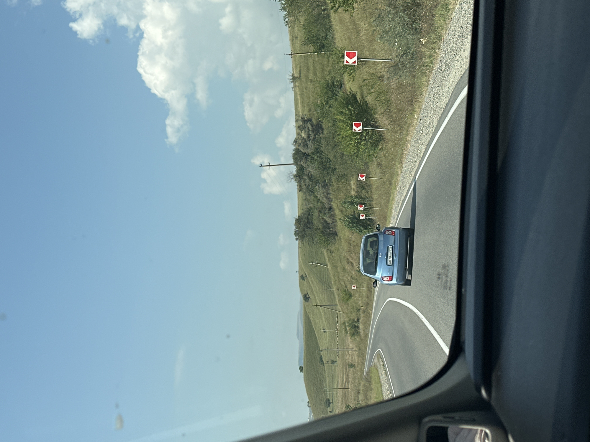 The image depicts a rural roadside scene, likely in or around Khabez, Russia, under a bright, partly cloudy sky. A blue hatchback is driving on a paved, winding road. The car has a license plate that reads "H878AO 777". The road is flanked by a gravel shoulder on the right and a grassy embankment on the left. Warning signs with red arrows indicating a curve are visible along the right side of the road. The landscape is characterized by rolling green hills dotted with trees and scattered utility poles. The scene is captured from inside another vehicle, with the dashboard and a portion of the interior visible in the foreground. The overall impression is one of a scenic drive through a hilly, rural area.