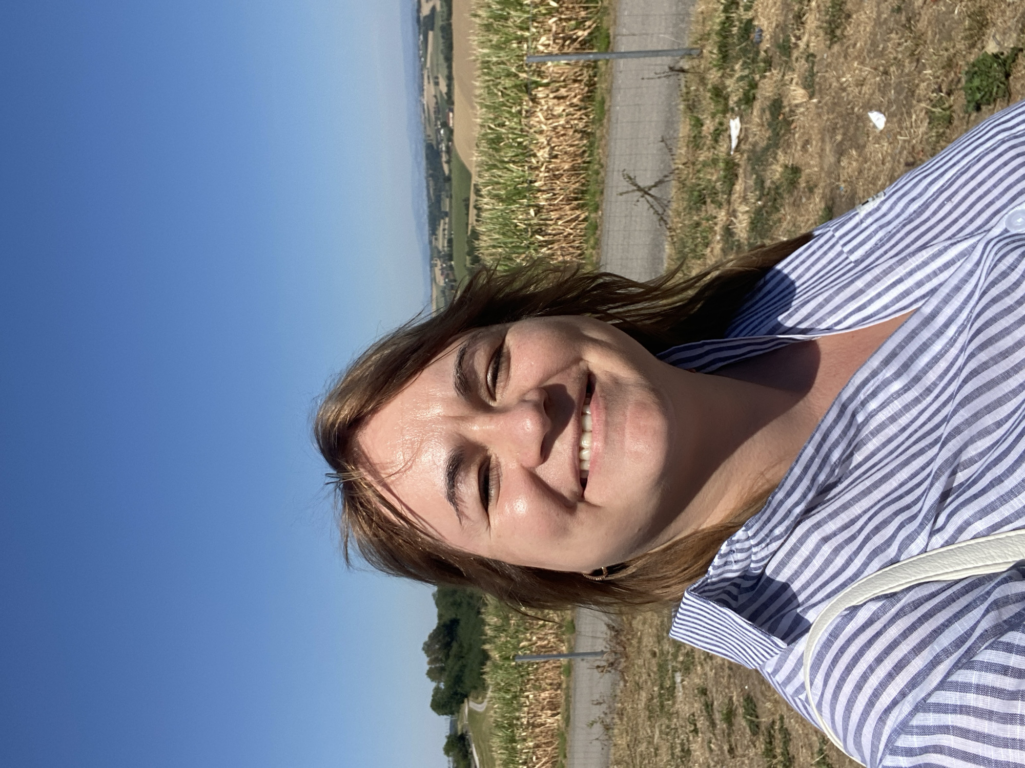 A woman with brown hair is taking a selfie outdoors on a sunny day. She is wearing a blue and white striped shirt and a white strap is visible over her shoulder. Behind her, there is a field of tall dry grass and corn stalks. In the distance, there are rolling hills with trees and some buildings. The sky is clear and blue. The image appears to be taken in a rural or agricultural area. The location is identified as Ramsau, Austria.