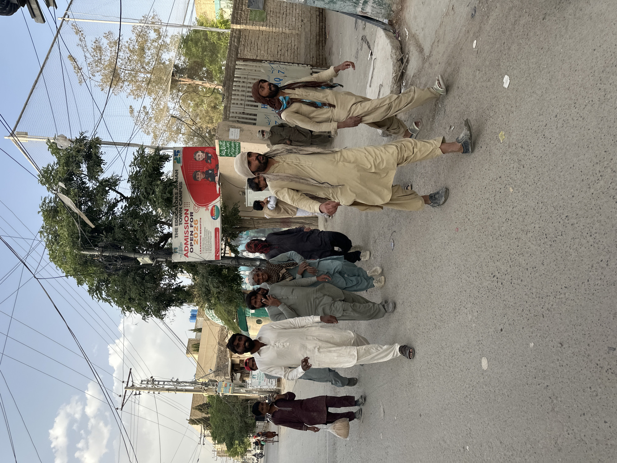 A group of seven men stand in a line along a paved street in Quetta City Tehsil, Pakistan. Most of the men are dressed in traditional Pakistani clothing, including shalwar kameez. The man at the front of the line, closest to the viewer, wears a dark maroon shalwar kameez and carries a white plastic bag. The second man wears a white shalwar kameez. The third man is on his phone. The fourth man is in a light grey shalwar kameez. The next man wears a dark grey shalwar kameez. The next two men are wearing lighter colored shalwar kameez with traditional head coverings. A building is on the right, and a tree and advertisement are on the left. Overhead is a mess of powerlines.