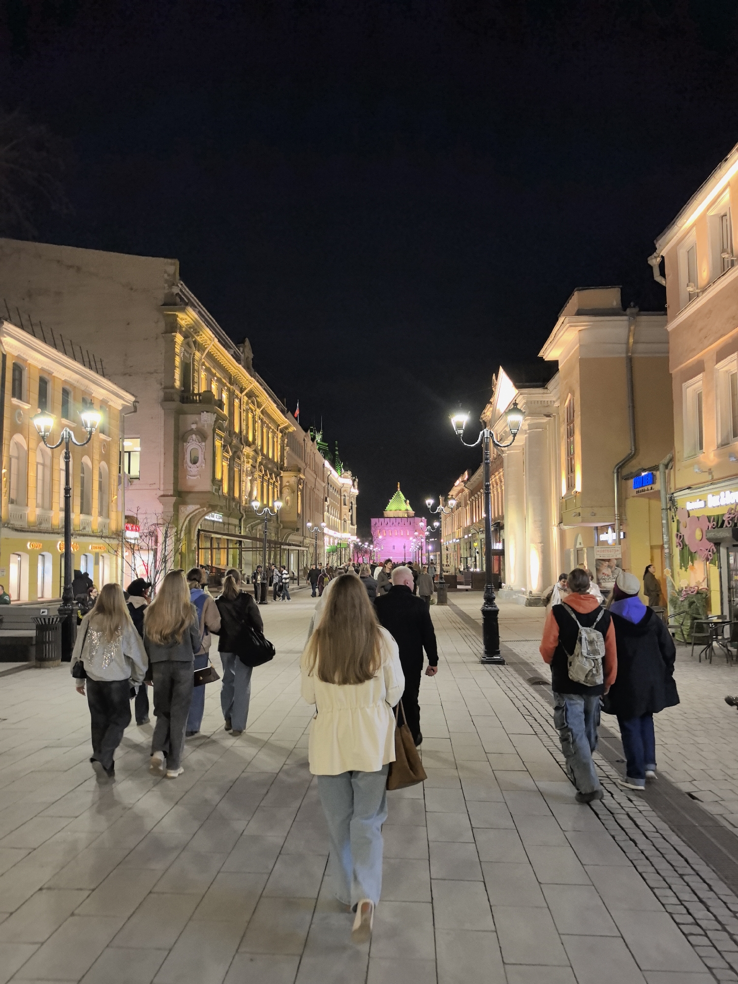 The image shows a nighttime view of a pedestrian street in Nizhny Novgorod, Russia. A group of people are walking away from the camera down the center of the paved street. The street is lined with illuminated buildings of varying architectural styles. Street lights are interspersed along the street. In the distance, a structure with a green dome is visible, likely part of the Nizhny Novgorod Kremlin. The sky above is dark.