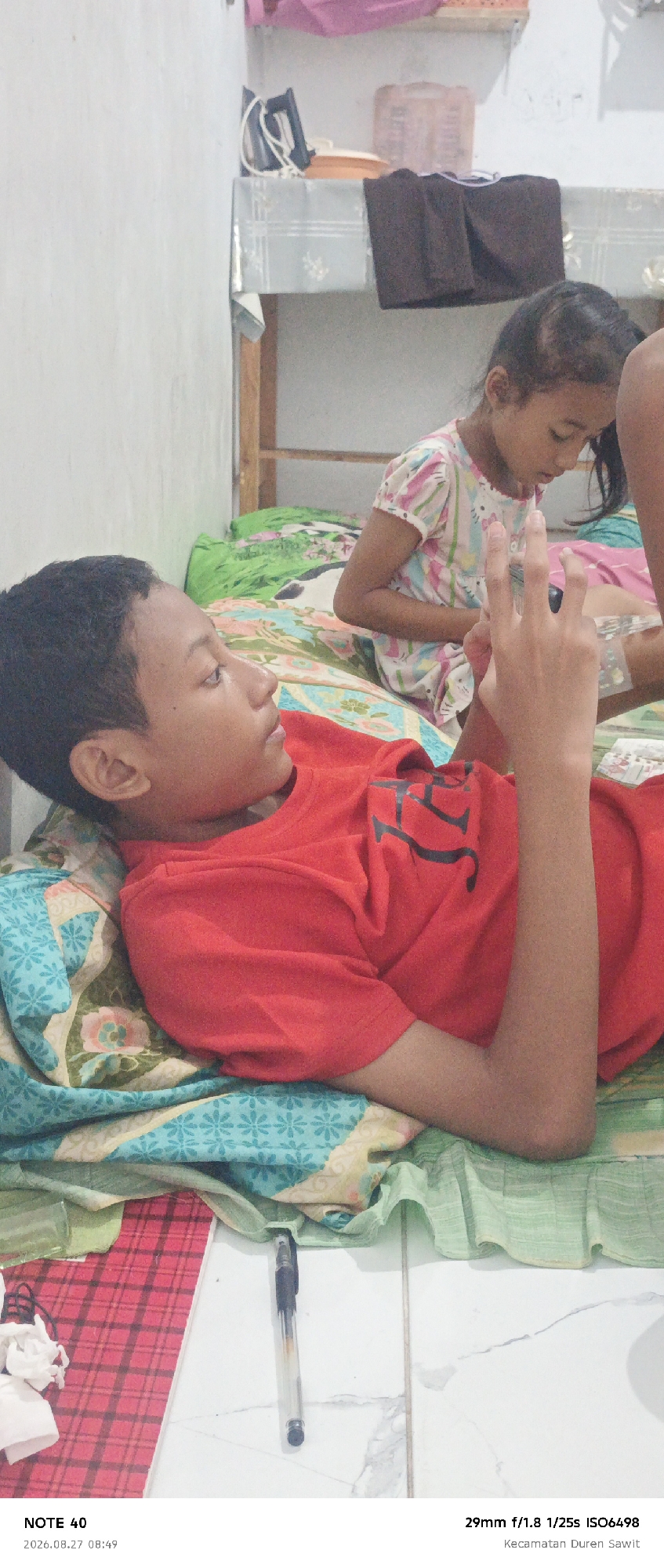A young male wearing a red shirt is lying on a bed with colorful patterned bedding. Next to him, a young female in a white and pink patterned dress is also on the bed. In the foreground, a pen lies on a red and white checkered notepad. Behind the children, a shelf with a black iron and a pink container is visible. The scene appears to be indoors, possibly in a bedroom. The text at the bottom of the image indicates the location as Kecamatan Duren Sawit, East Jakarta, Indonesia, and the timestamp as 2026.08.27 08:49.
