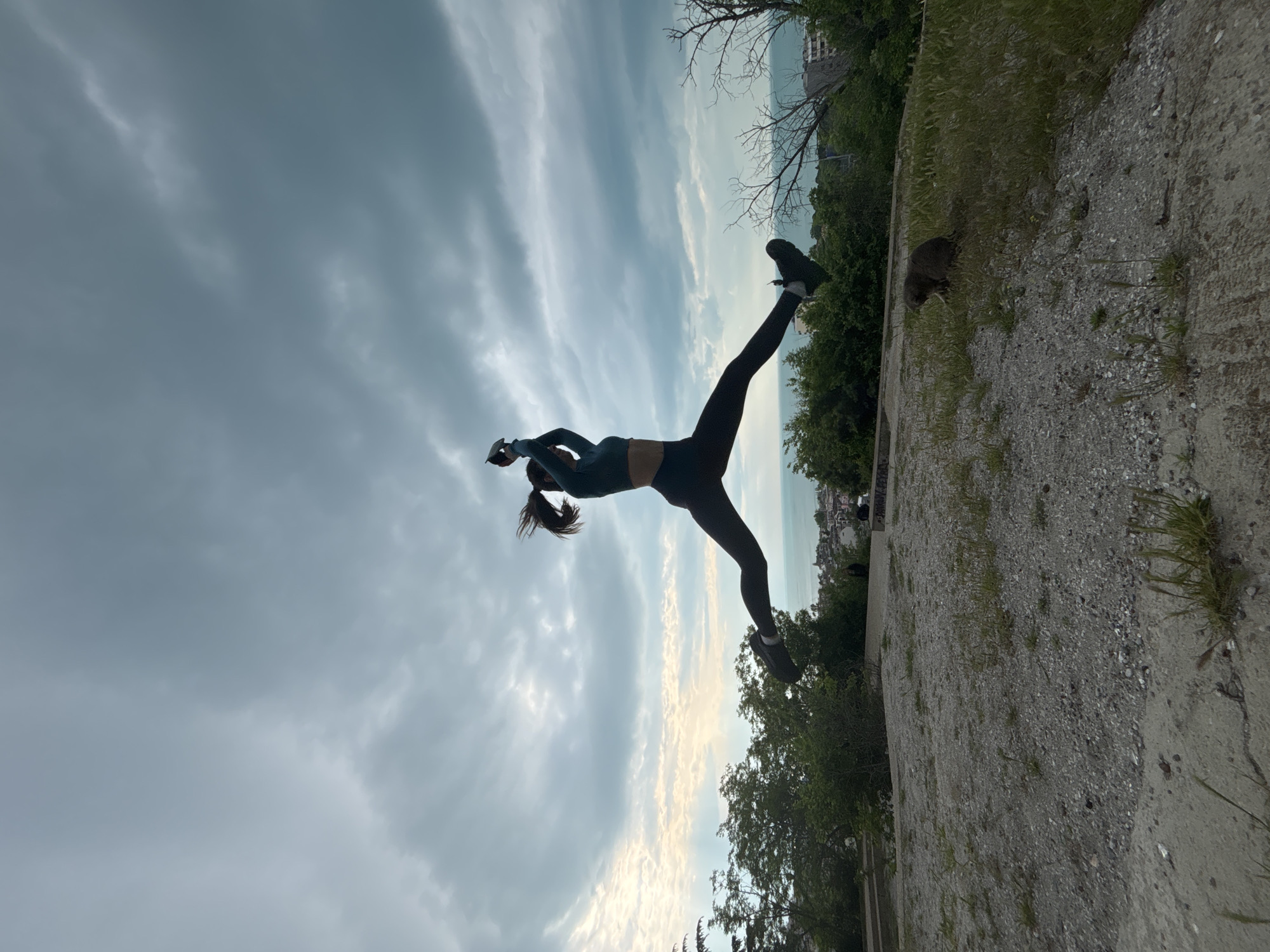 A woman in athletic wear is performing a split jump with one leg extended up toward the sky and the other angled down towards a stone ground. She is silhouetted against a cloudy sky. The setting appears to be an outdoor urban environment with trees and a glimpse of the coastline in the background. There is a rodent on the ground near the edge of the stone path. The location is most likely in the city of Varna, Bulgaria.